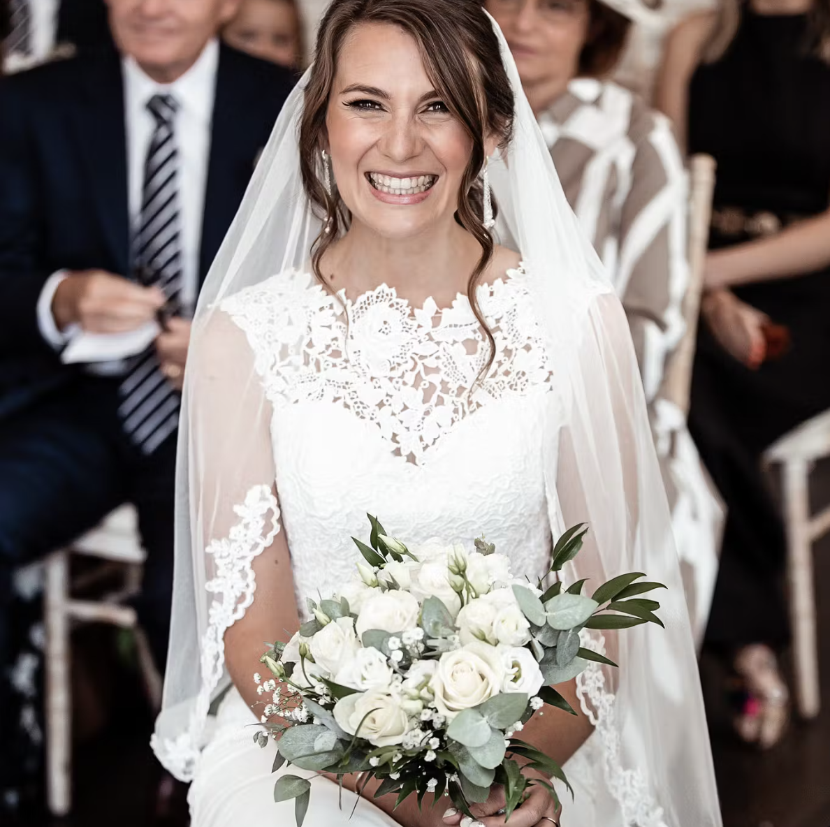 A bride in a white lace wedding dress holding a bouquet of white roses and greenery, smiling at the camera during a wedding ceremony.