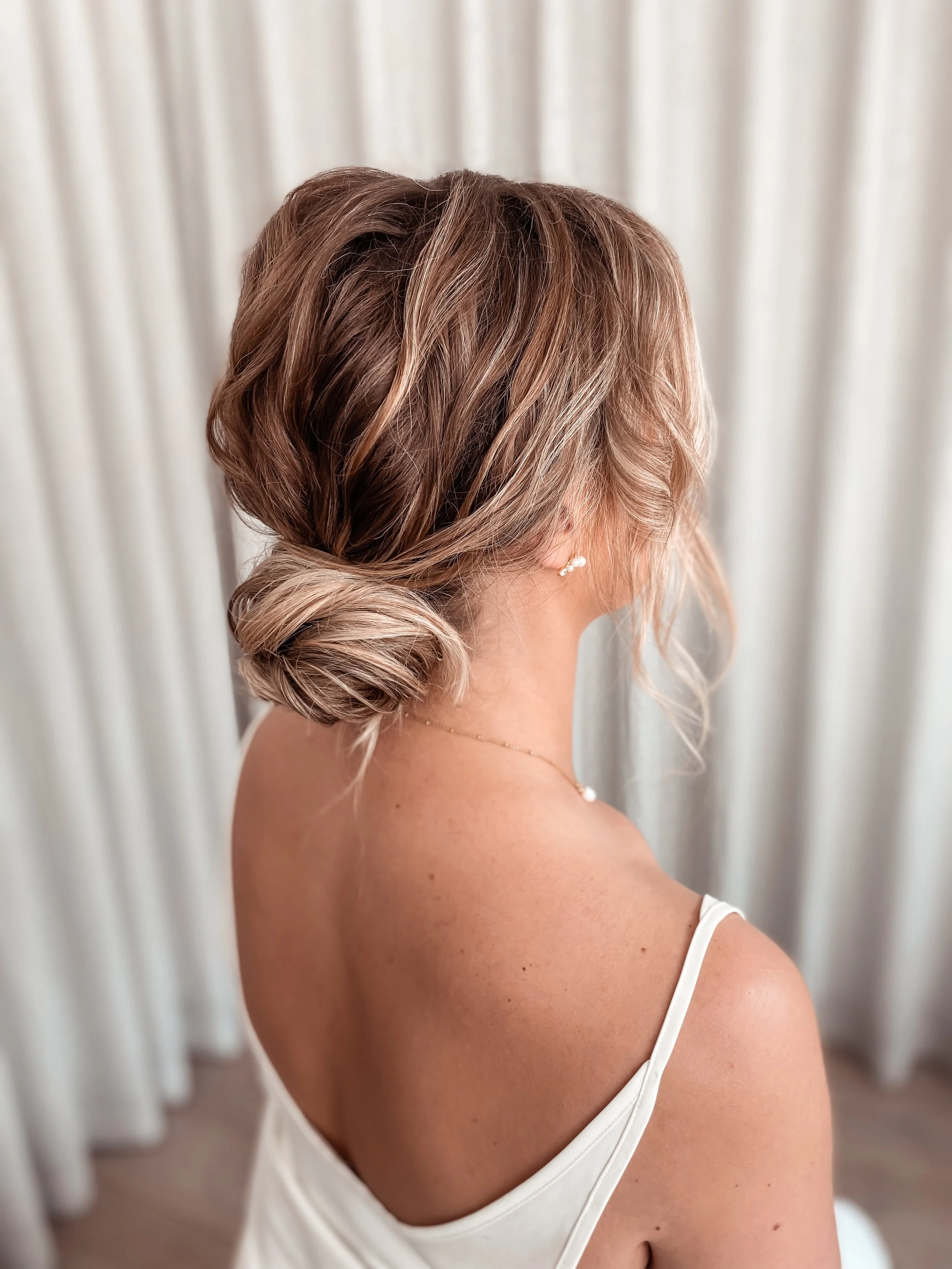 Side view of a woman with styled light brown hair in a loose bun, wearing a white top with thin straps, pearl earrings, and a delicate necklace, standing in front of white curtains.