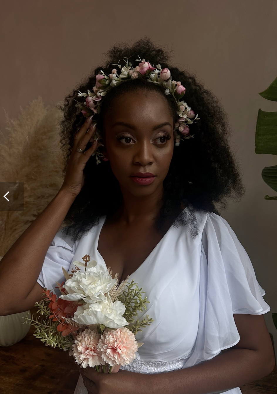 A woman with dark, curly hair wearing a floral crown and a white dress holding a bouquet of white and pink flowers.
