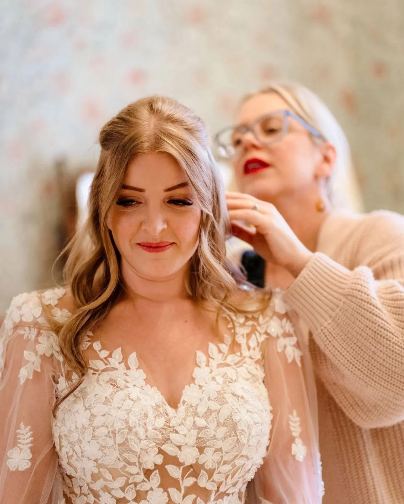 A woman with light skin and wavy blonde hair smiling softly while a makeup artist, older woman with glasses, is applying makeup to her face.