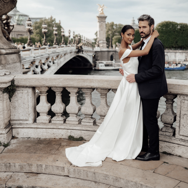 A newlywed couple in elegant wedding attire standing on a stone bridge by a river, with historic architecture and a statue in the background.