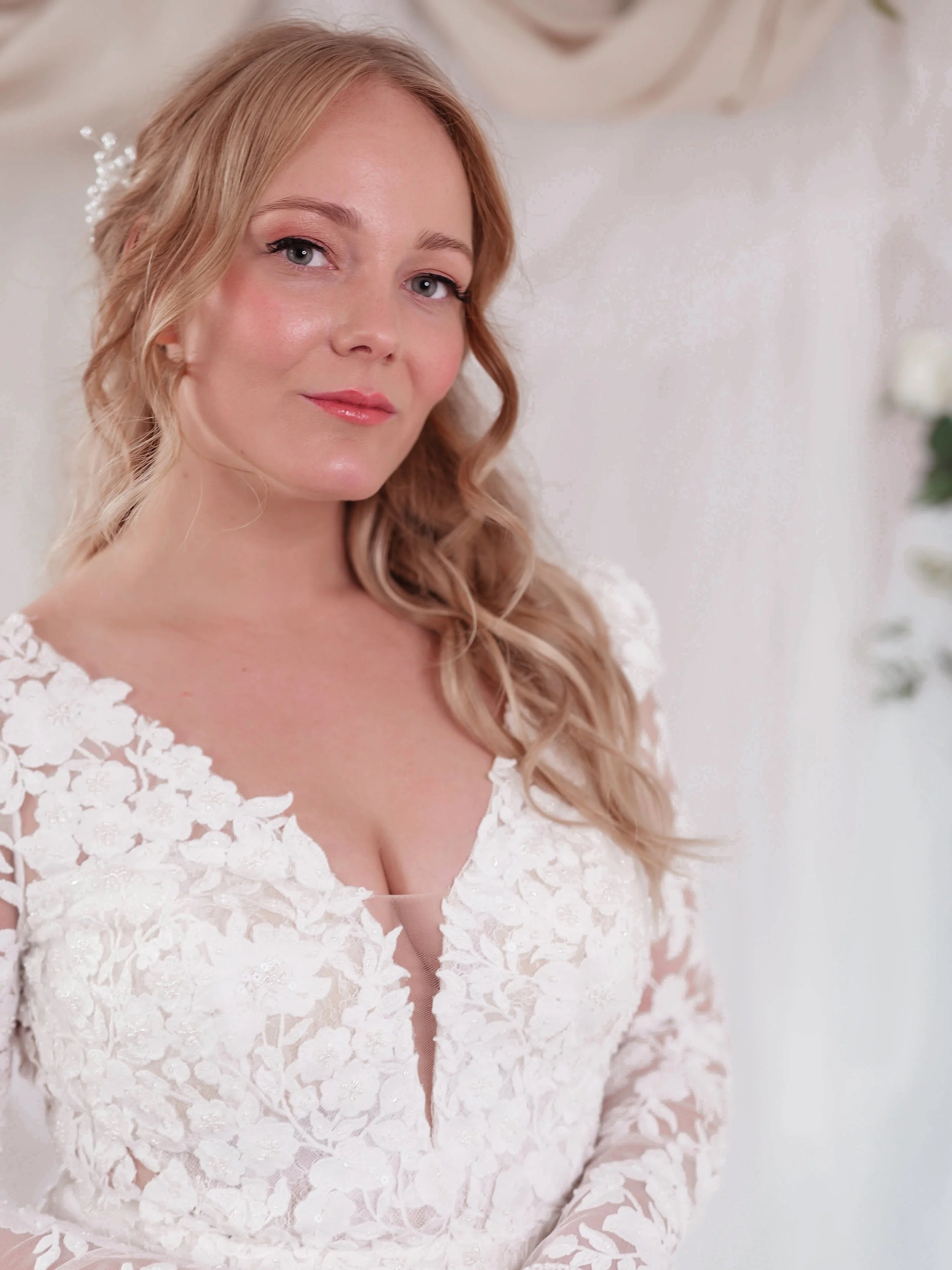 A woman with long, wavy blonde hair wearing a white lace dress, standing indoors with a soft background, possibly at a wedding or special event.