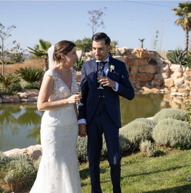 Bride and groom standing outdoors near a pond, holding hands and glasses of champagne during their wedding celebration.