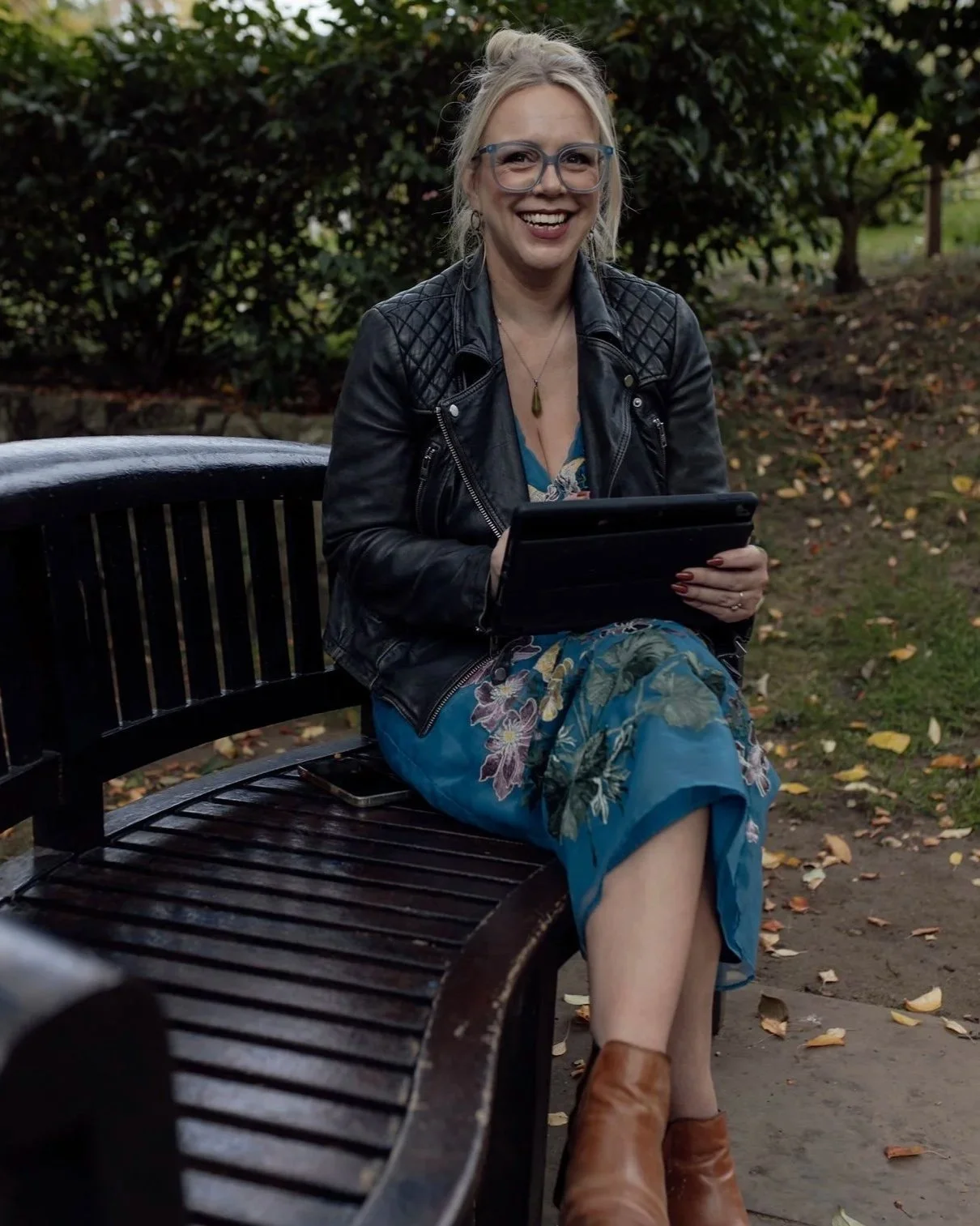 A woman sitting on a park bench, smiling and holding an electronic device, wearing glasses, a black leather jacket, a blue floral dress, and brown boots, with greenery and fallen leaves in the background.