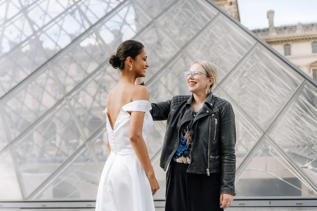 Two women smiling and laughing in front of the Louvre Pyramid in Paris, one in a white dress and the other in a black leather jacket.