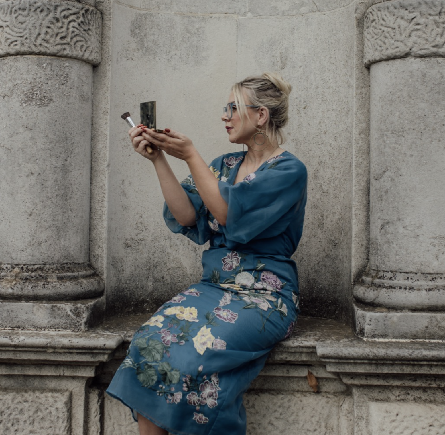 A woman in a blue floral dress with glasses and earrings sitting on a stone ledge in front of an ornate stone wall, holding a makeup brush and compact mirror, looking at herself.