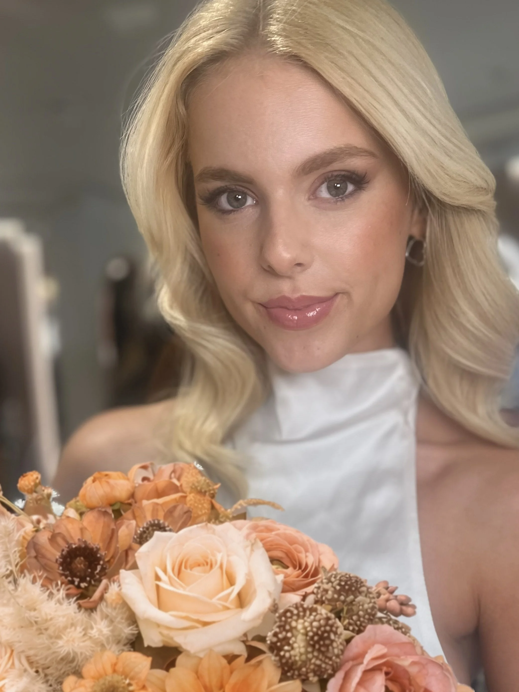 A woman with blonde hair and light makeup holding a bouquet of peach and orange flowers, smiling indoors.
