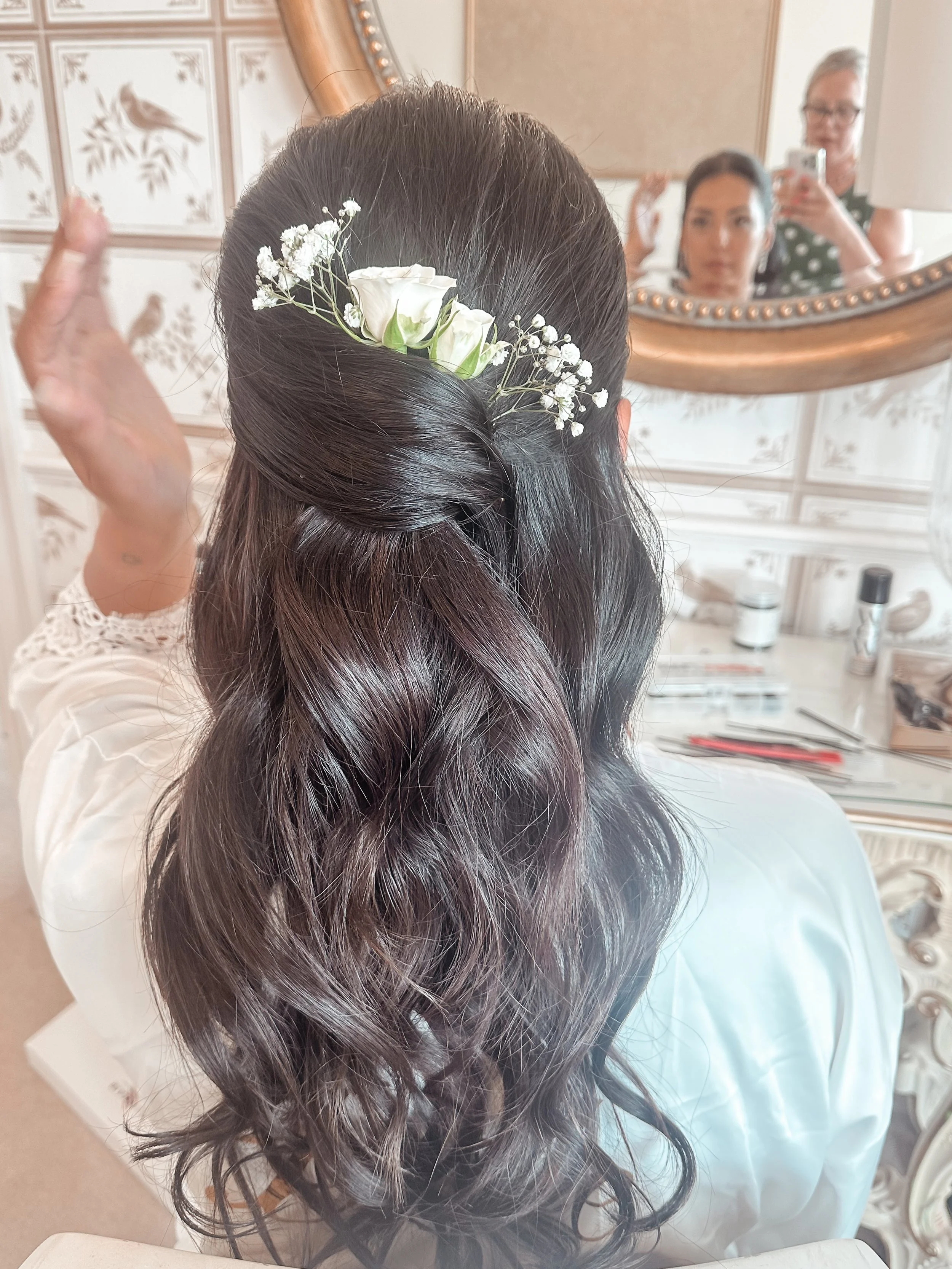 Back of a woman's head showing long wavy dark hair adorned with white flowers, in front of a mirror where two women are taking a photo.