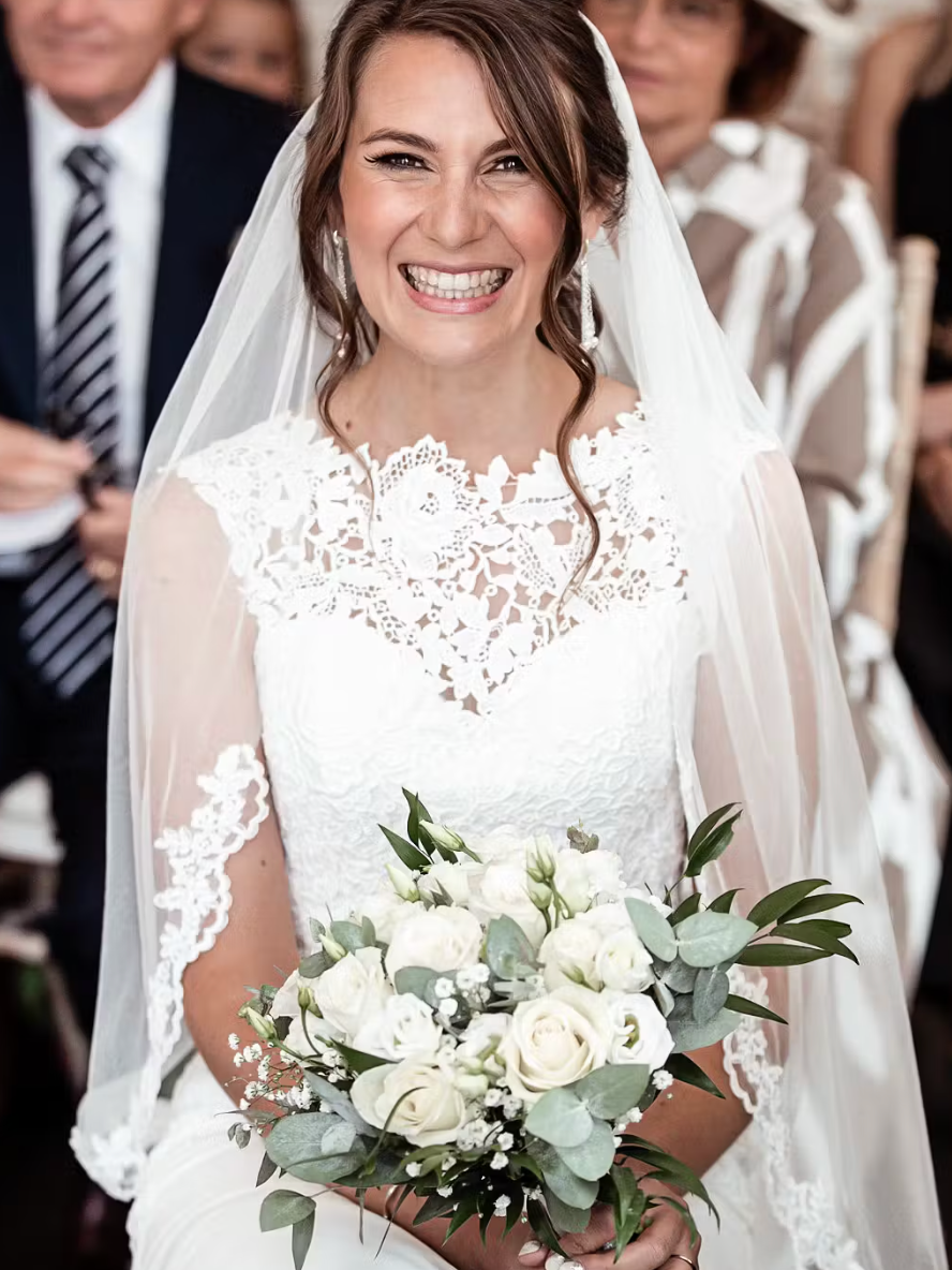 A bride in a white lace wedding dress holding a bouquet of white roses and greenery smiles at the camera. She wears a veil, and guests are seated in the background.