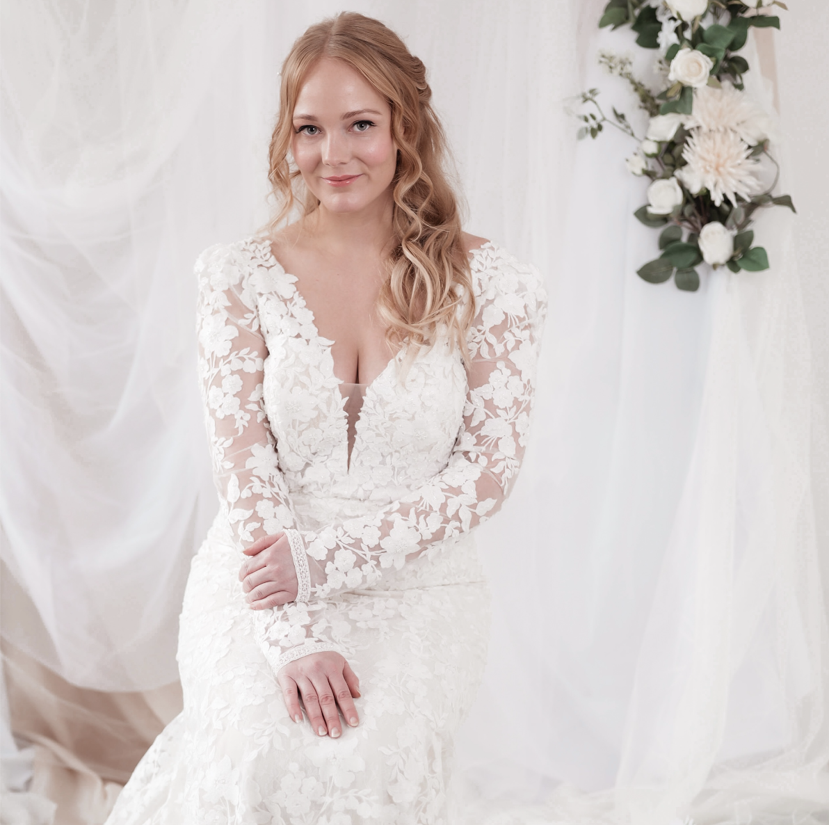 A woman in a white lace wedding dress sitting with her hands on her lap, smiling softly, with a floral arrangement in the background.