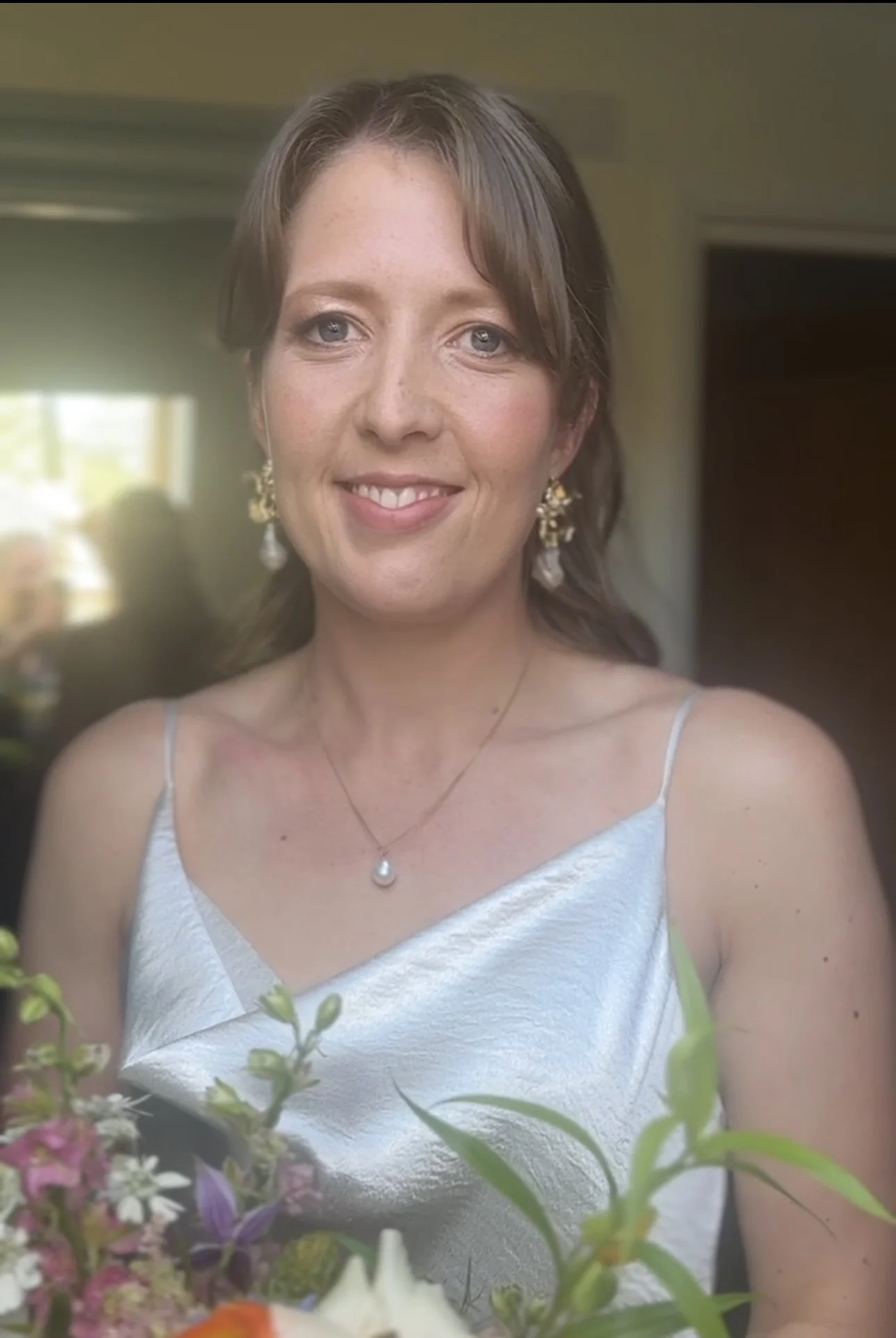 A woman with shoulder-length brown hair, blue eyes, wearing a white silk sleeveless dress and gold jewelry, smiling indoors with natural light coming from a window.