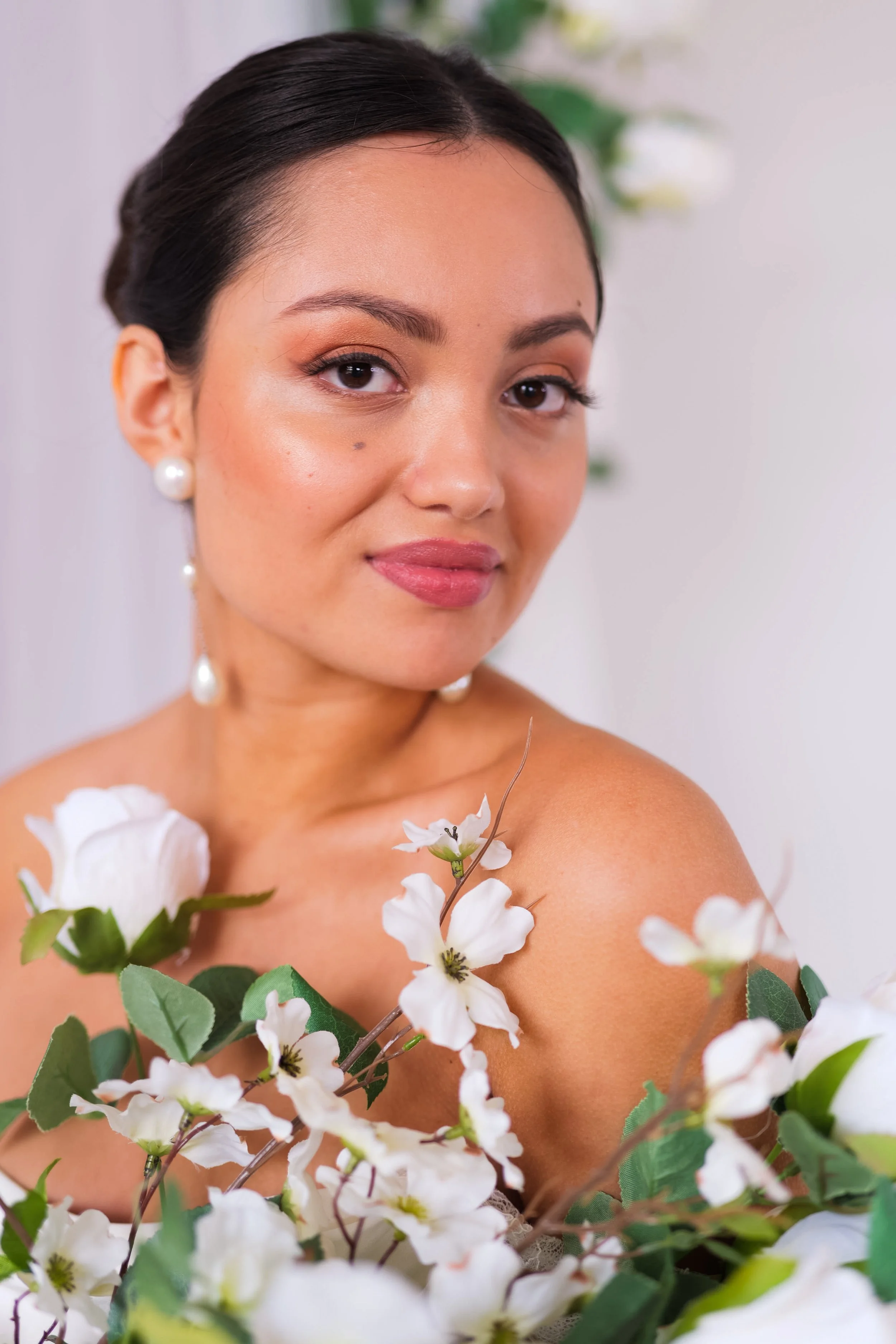 Close-up of a woman with dark hair in an elegant hairstyle, wearing pearl earrings, surrounded by white flowers, looking at the camera with a slight smile.