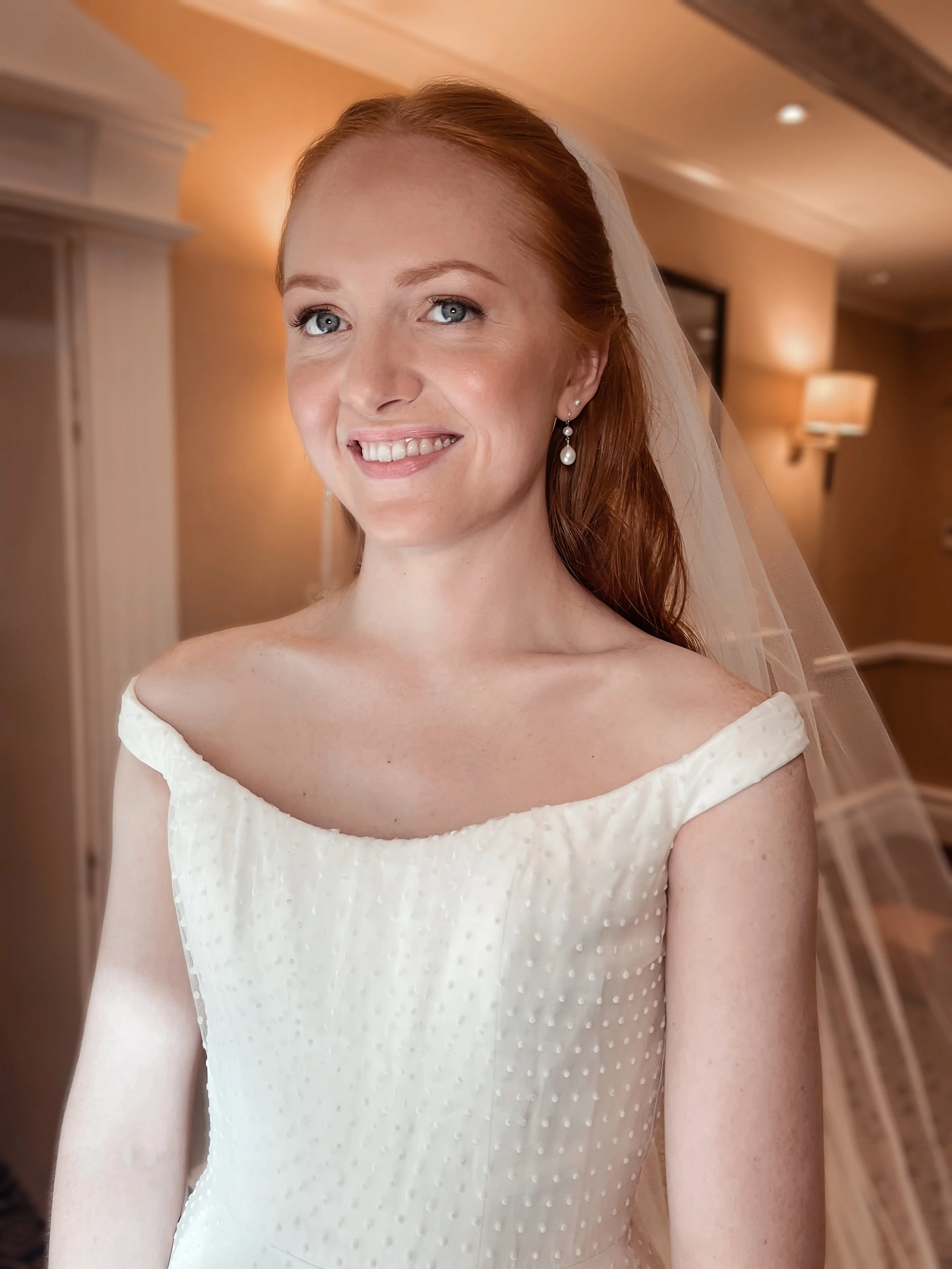 A smiling bride with red hair, blue eyes, wearing pearl earrings and a white wedding dress with off-the-shoulder sleeves and a sheer veil, standing indoors.