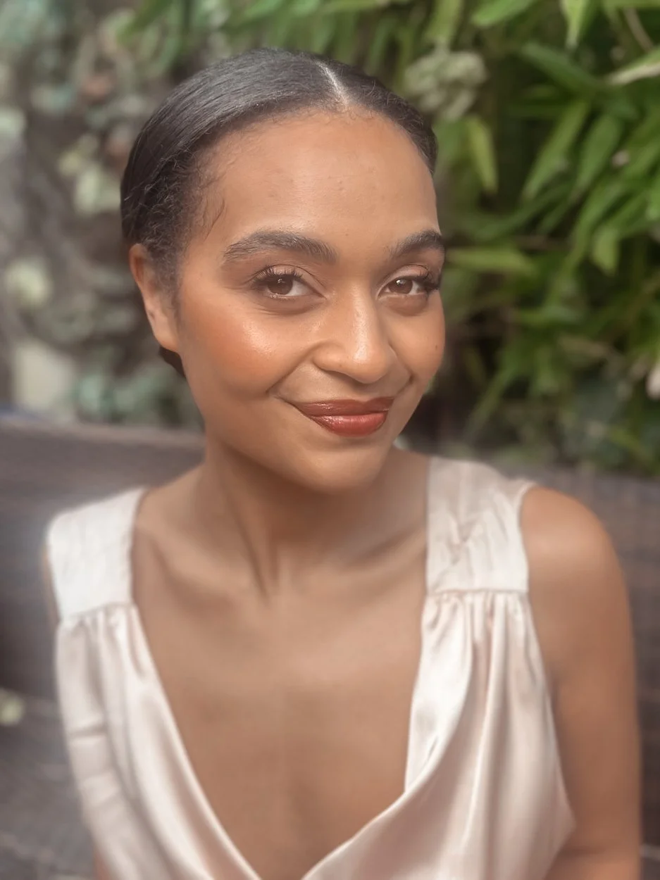 A woman with smooth skin and short dark hair, smiling softly, wearing a cream-colored satin sleeveless top, with green foliage in the background.