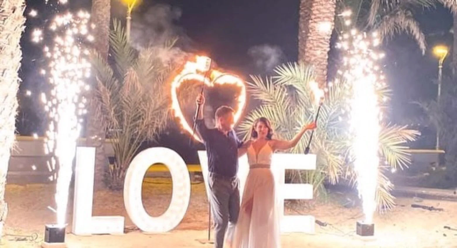 A couple celebrating love at night with sparklers in front of large illuminated letters spelling 'LOVE', with palm trees and sparklers creating a festive atmosphere.