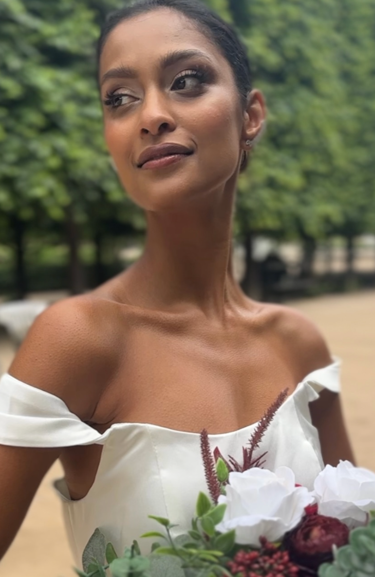 Woman with dark hair in an off-shoulder white dress holding a bouquet of flowers outdoors.