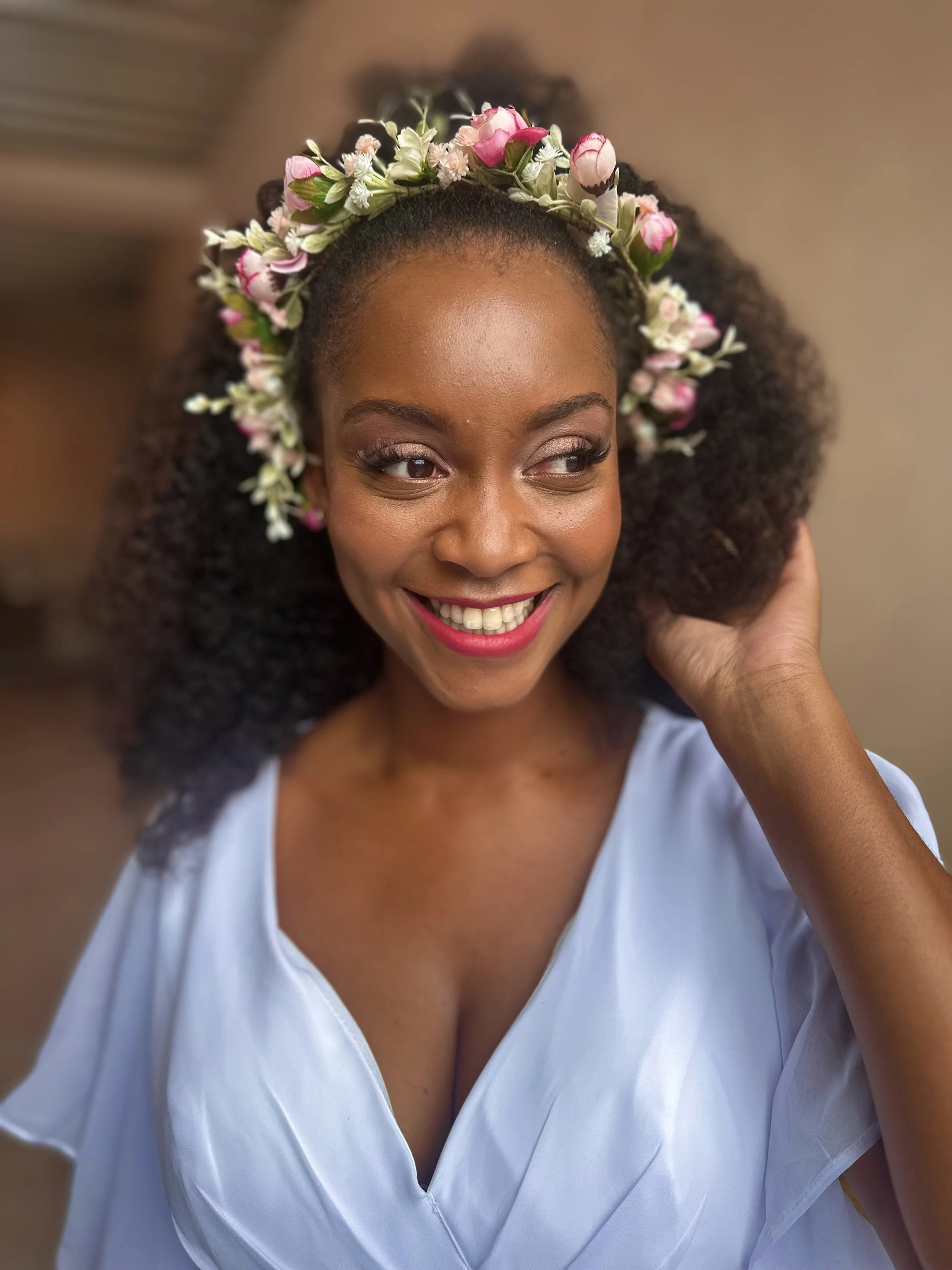 A smiling woman wearing a floral crown and a white top, touching her hair.