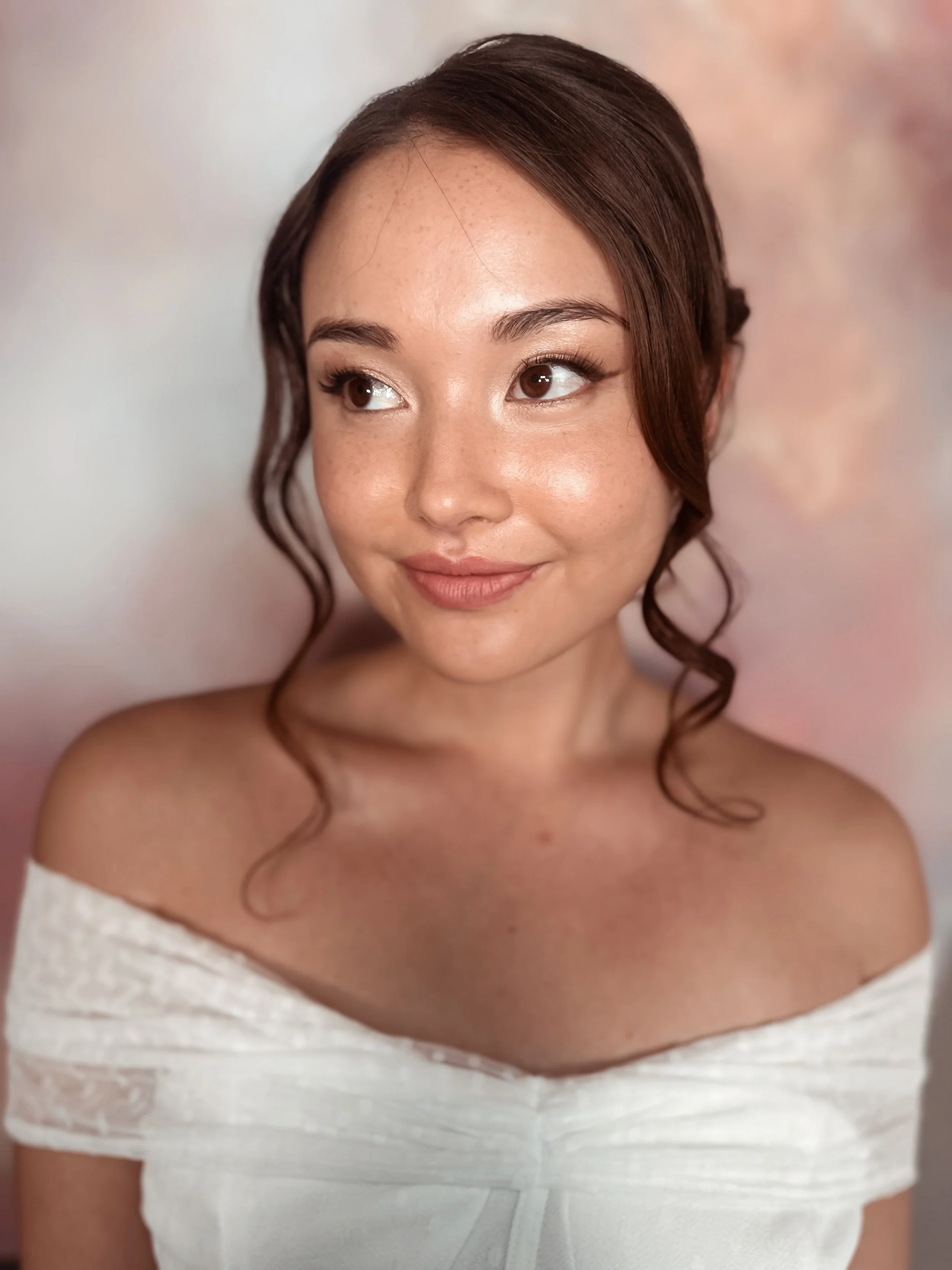 A young woman with brown hair styled in loose curls, wearing an off-shoulder white dress, looking to her right with a soft smile against a pastel-colored background.