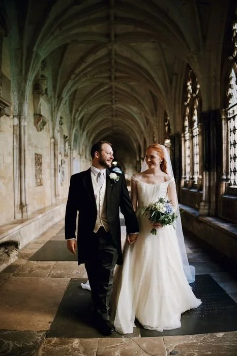 A newlywed couple walking hand in hand through Westminster abbey corridor with gothic arches and stained glass windows, celebrating their wedding day.