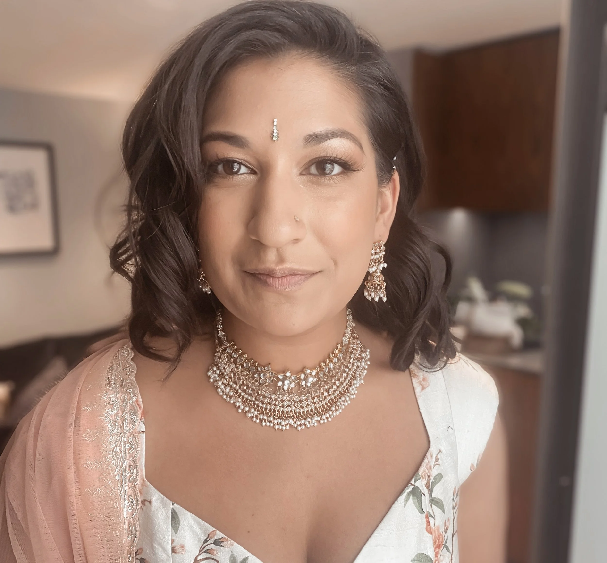 A woman with dark wavy hair, wearing traditional Indian jewelry including earrings, a necklace, and a bindi, smiling softly in an indoor setting.
