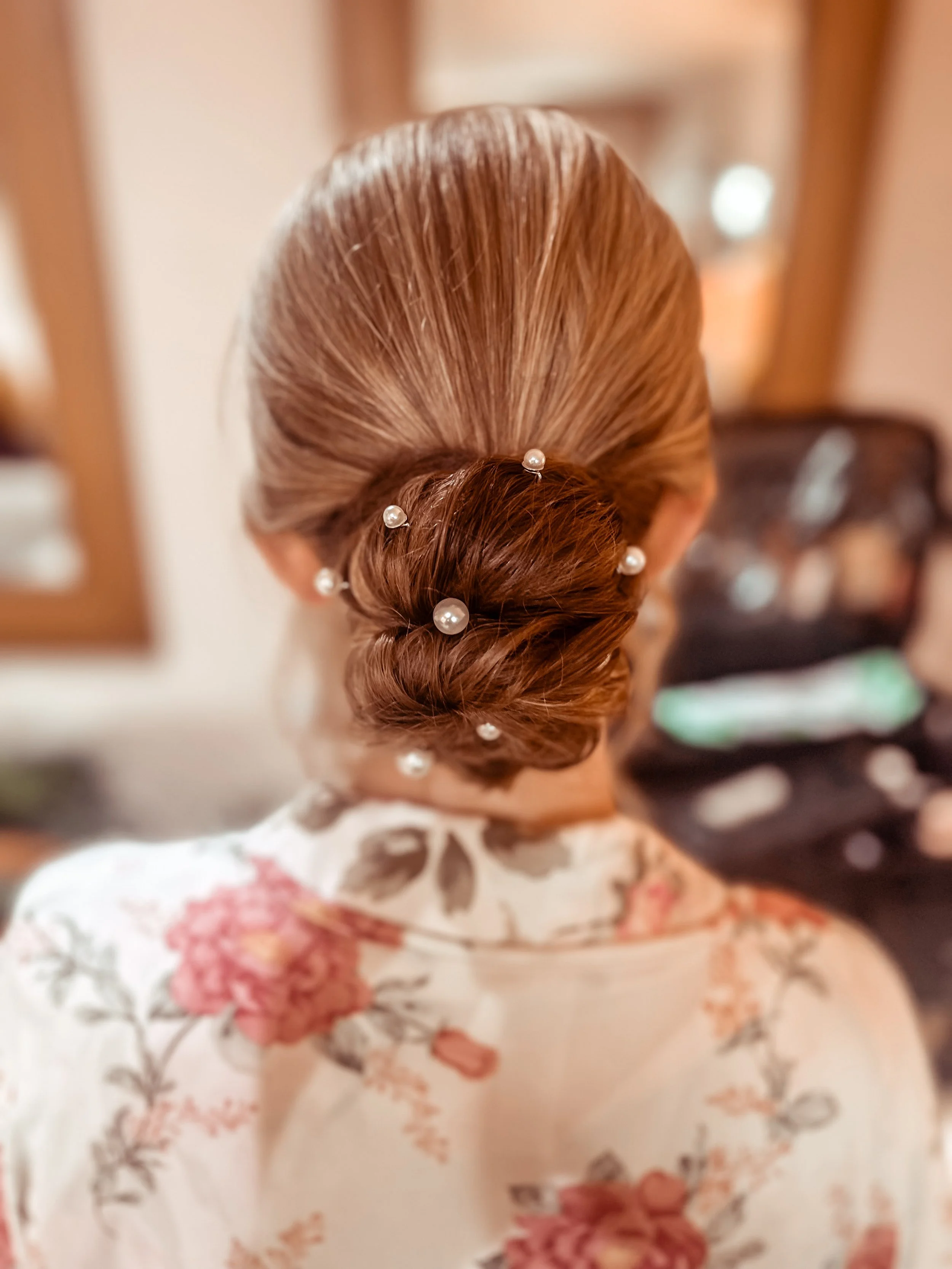 Back view of a woman with styled brown hair adorned with pearl pins, wearing a floral top, in a room with a mirror and blurred furniture in the background.