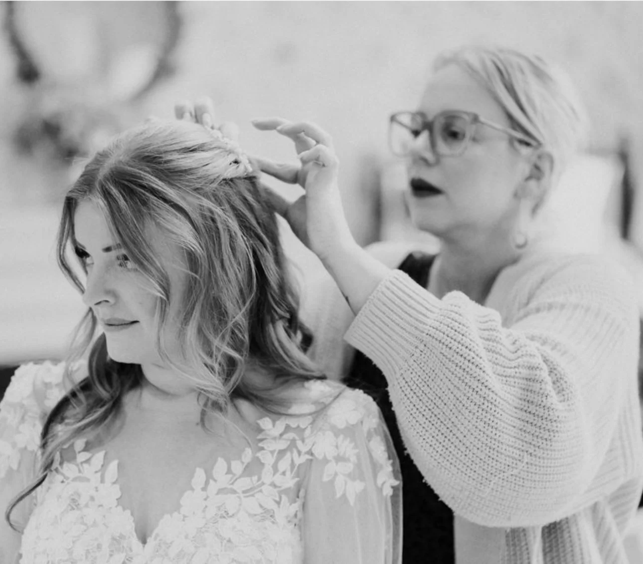 A woman with long, wavy hair looking down while a stylist touches her hair, preparing her for a special occasion.