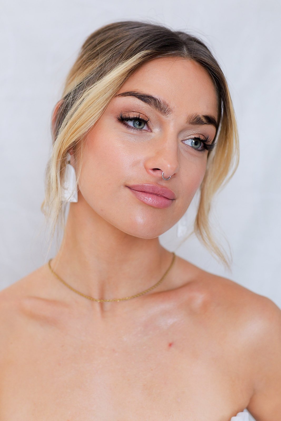 A young woman with blonde hair, blue eyes, and fair skin. She is wearing makeup, a gold necklace, and white earrings, and has a septum piercing. She is looking slightly to the side with a neutral expression.