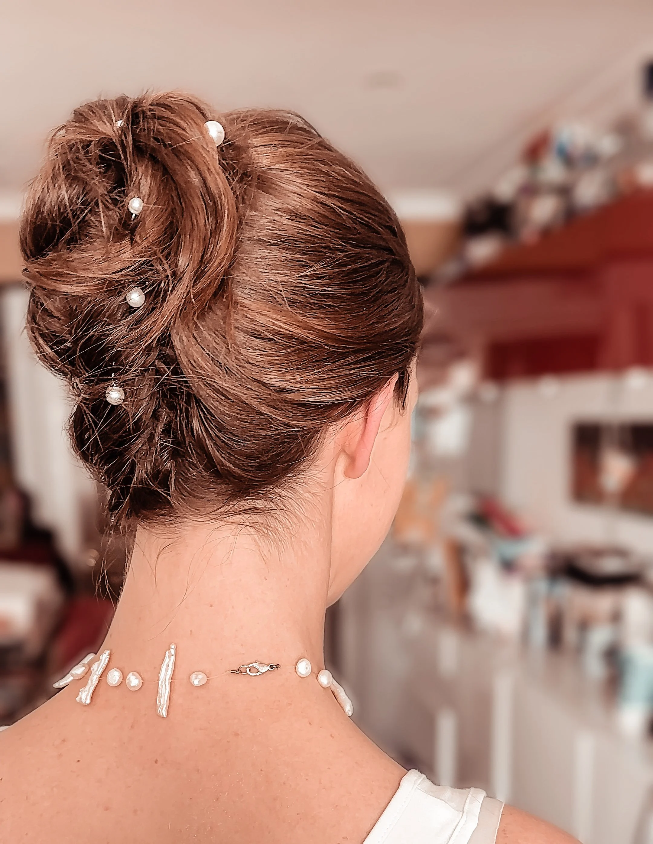 Side view of a woman with styled updo hair adorned with pearls. She is wearing a pearl necklace and a white top, with a blurred background of a kitchen.