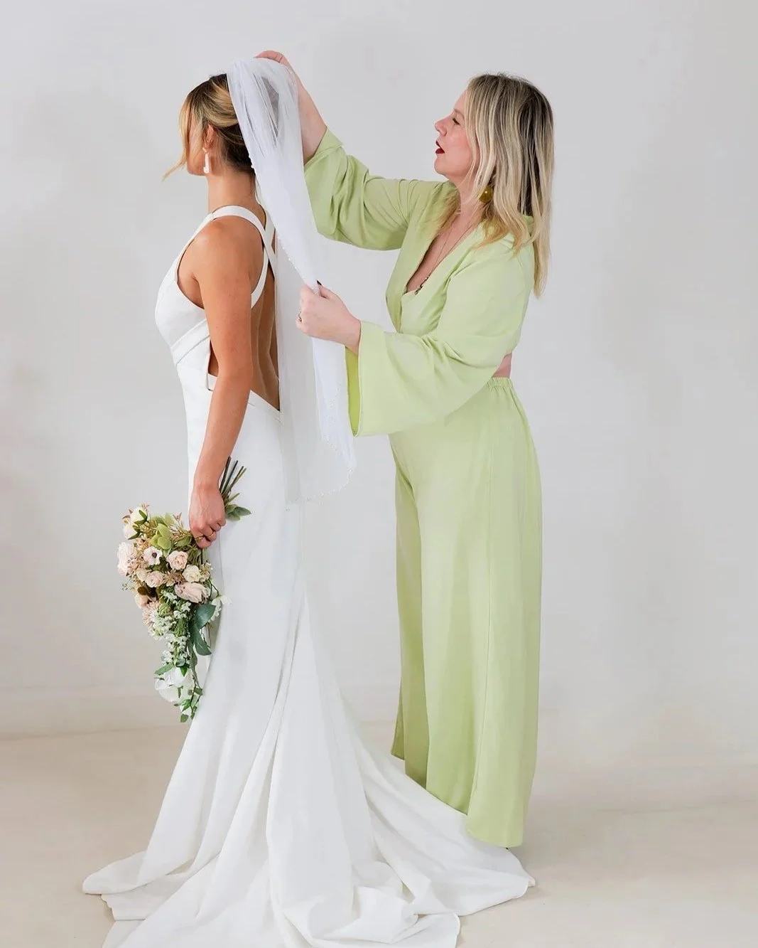 A woman in a white wedding dress holding a bouquet receives assistance from another woman in a lime green outfit as she adjusts her veil.