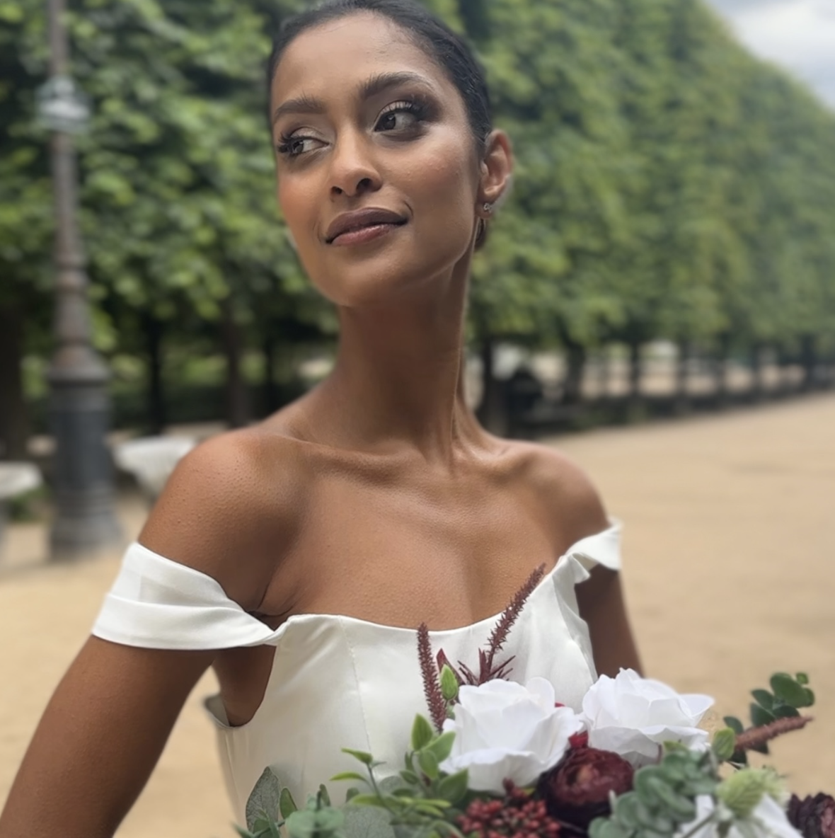 Woman in a white off-shoulder dress holding a bouquet of white and dark red flowers outdoors, with green trees in the background.