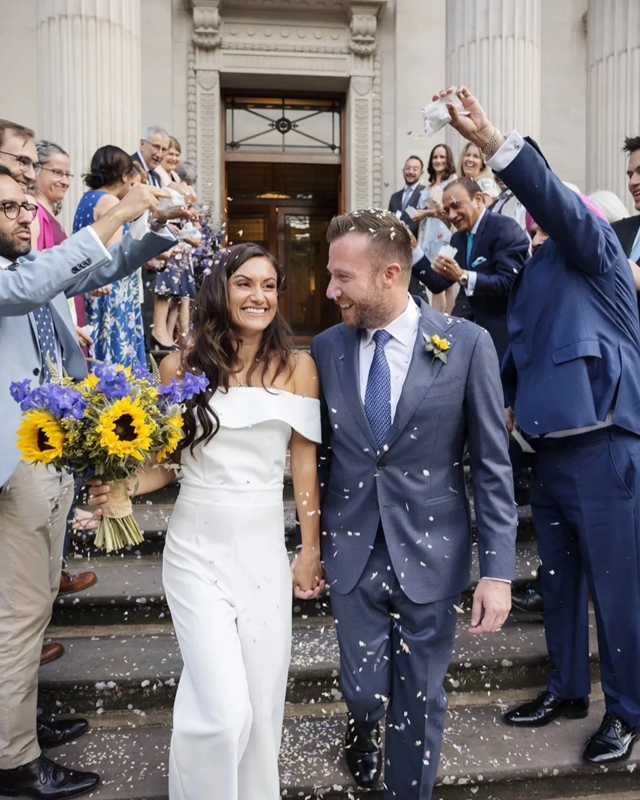 A newly married couple walking down the steps of a building, holding hands, with guests celebrating and throwing confetti around them.