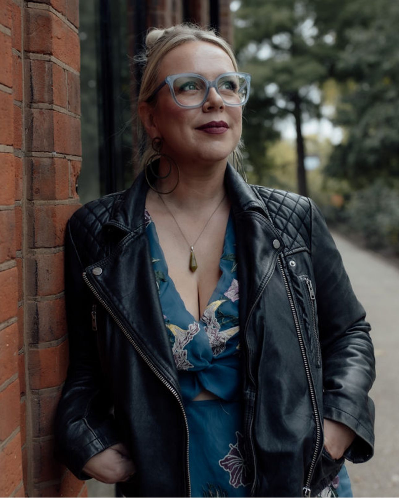 A woman with blonde hair pulled back, wearing glasses, a black leather jacket, and a blue floral dress, leaning against a brick wall outdoors on a cloudy day.