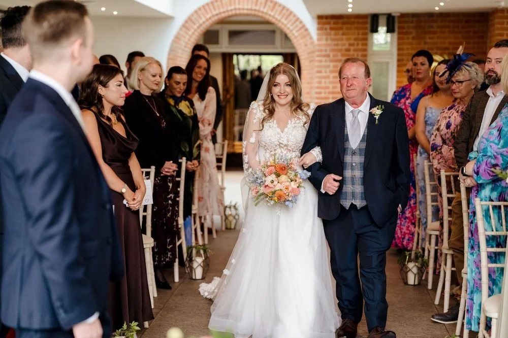 A bride walking down the aisle, escorted by an older man, at a wedding ceremony. Guests are standing on either side, watching and smiling.
