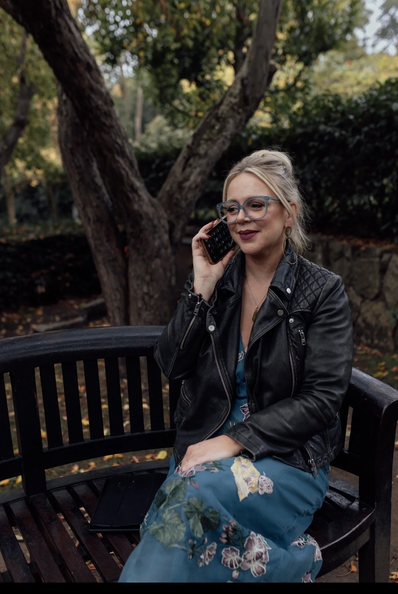 A woman with blonde hair, glasses, and earrings sitting on a black wooden park bench talking on a cell phone. She is wearing a black leather jacket and a blue floral dress, with a dark tablet on her lap. The background shows trees and bushes in an outdoor park setting.