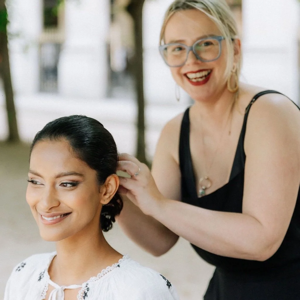 A woman with blonde hair, wearing glasses and a black dress, is smiling while styling a young woman's hair outdoors.