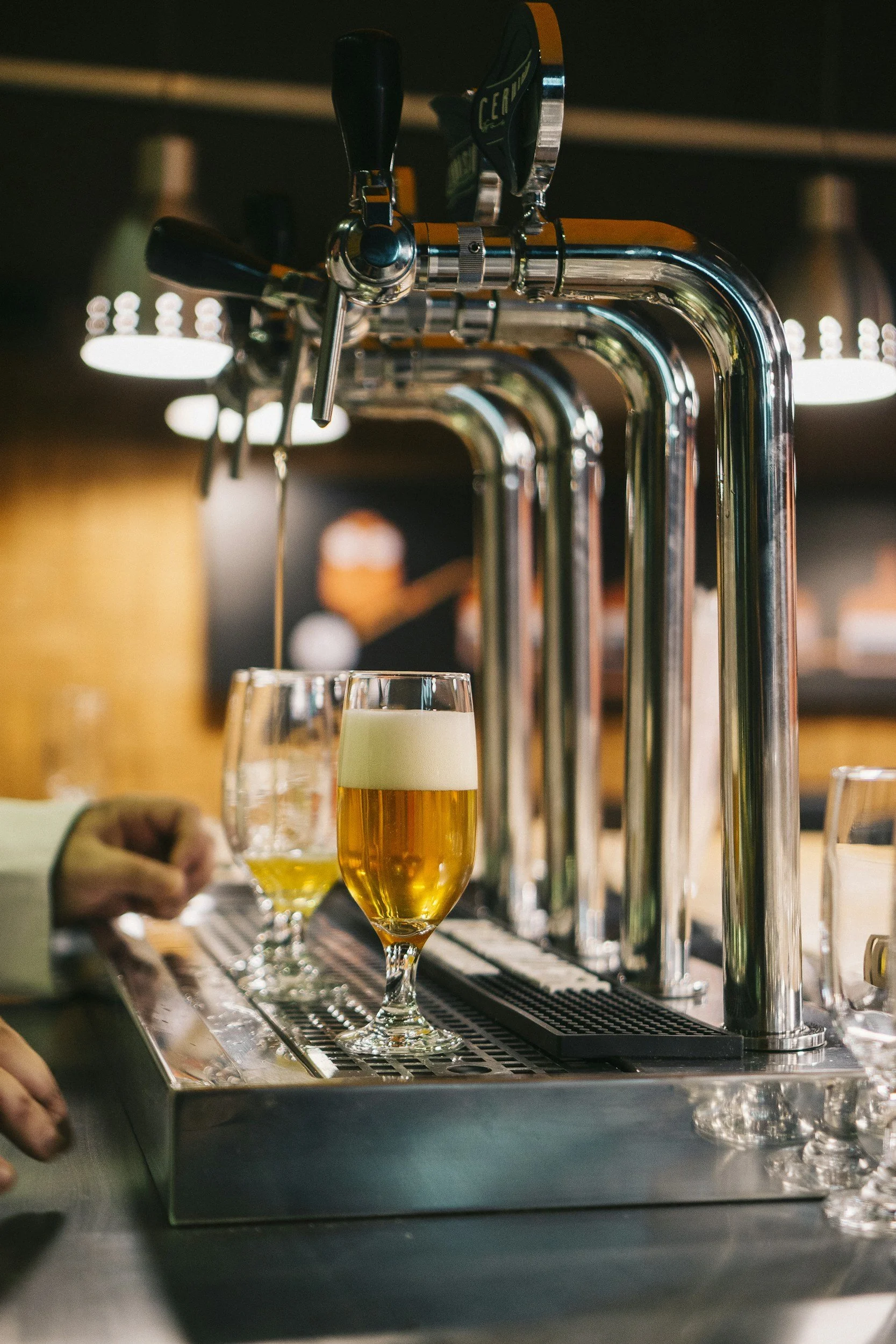 A glass of beer with foam being poured from a tap at a bar, with other glasses and a person’s hand nearby.