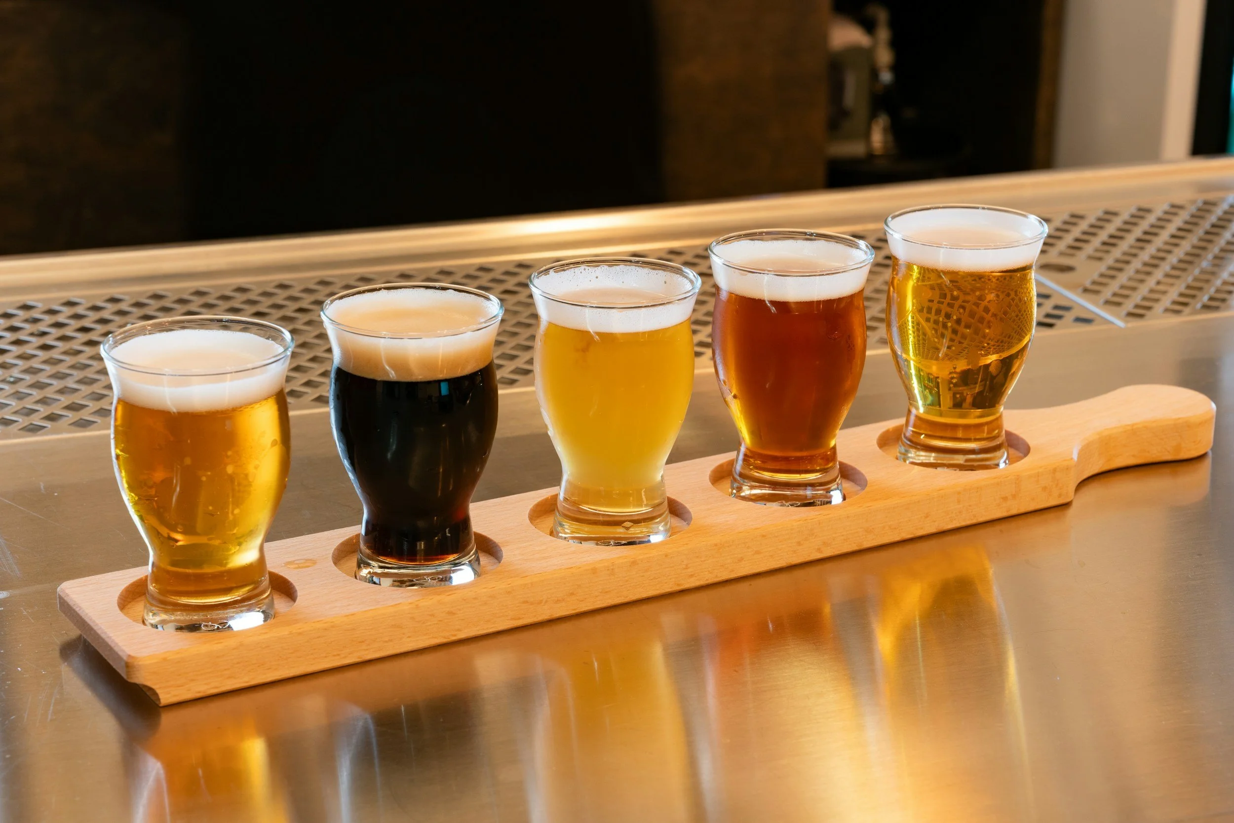 Five different types of beer in small glasses on a wooden tray on a bar counter.
