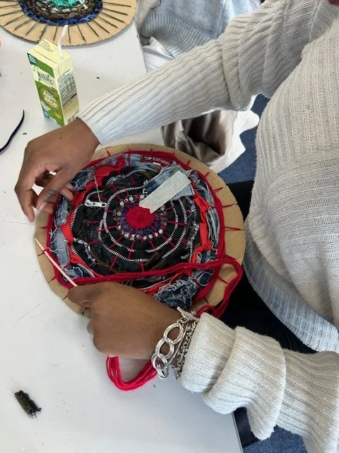Person working on a circular fabric piece, possibly a craft or embroidery project, at a table with a carton of juice nearby.