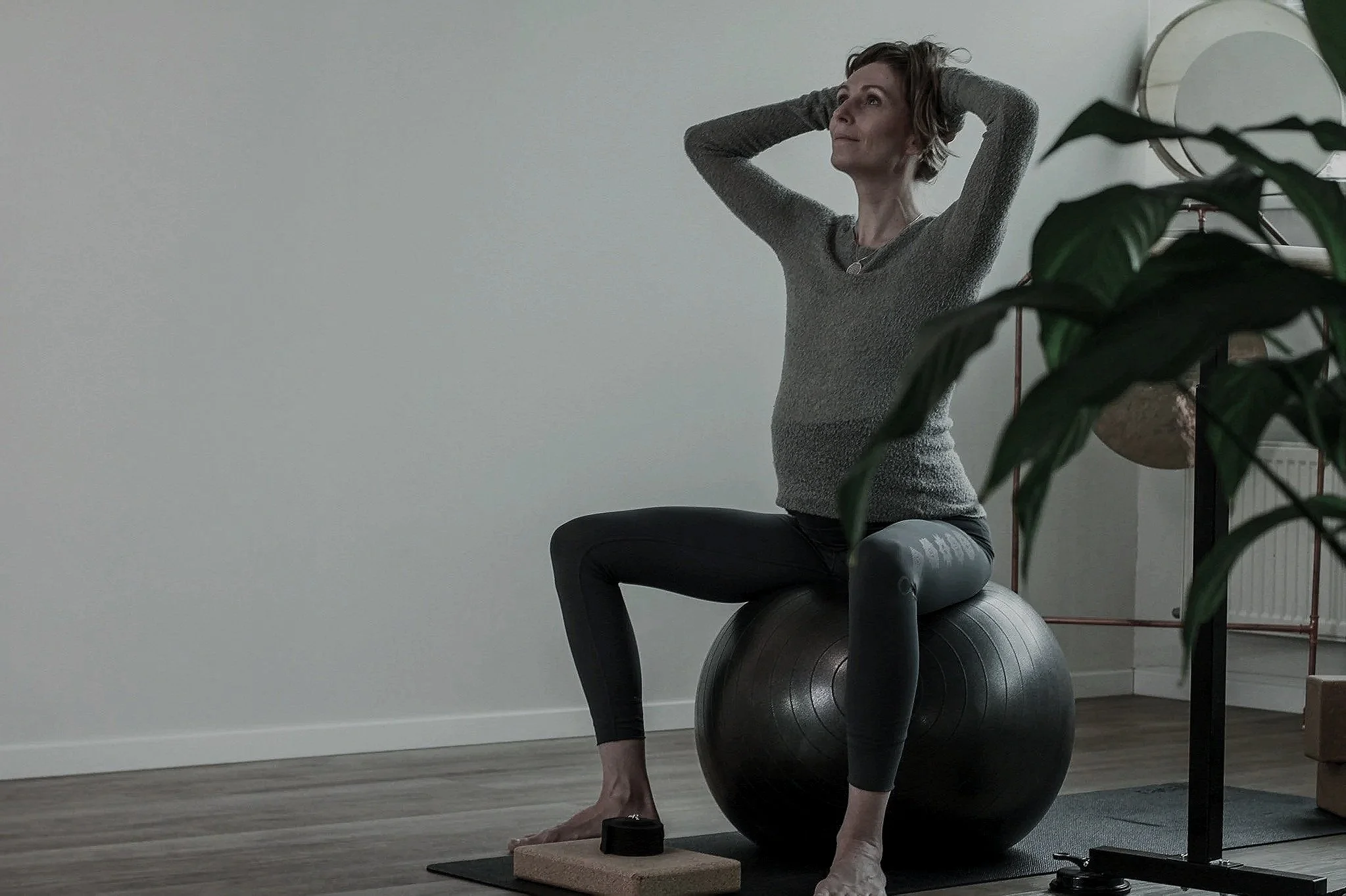 Woman practicing yoga or meditation while seated on a large exercise ball in a minimally decorated room, with her hands behind her head.