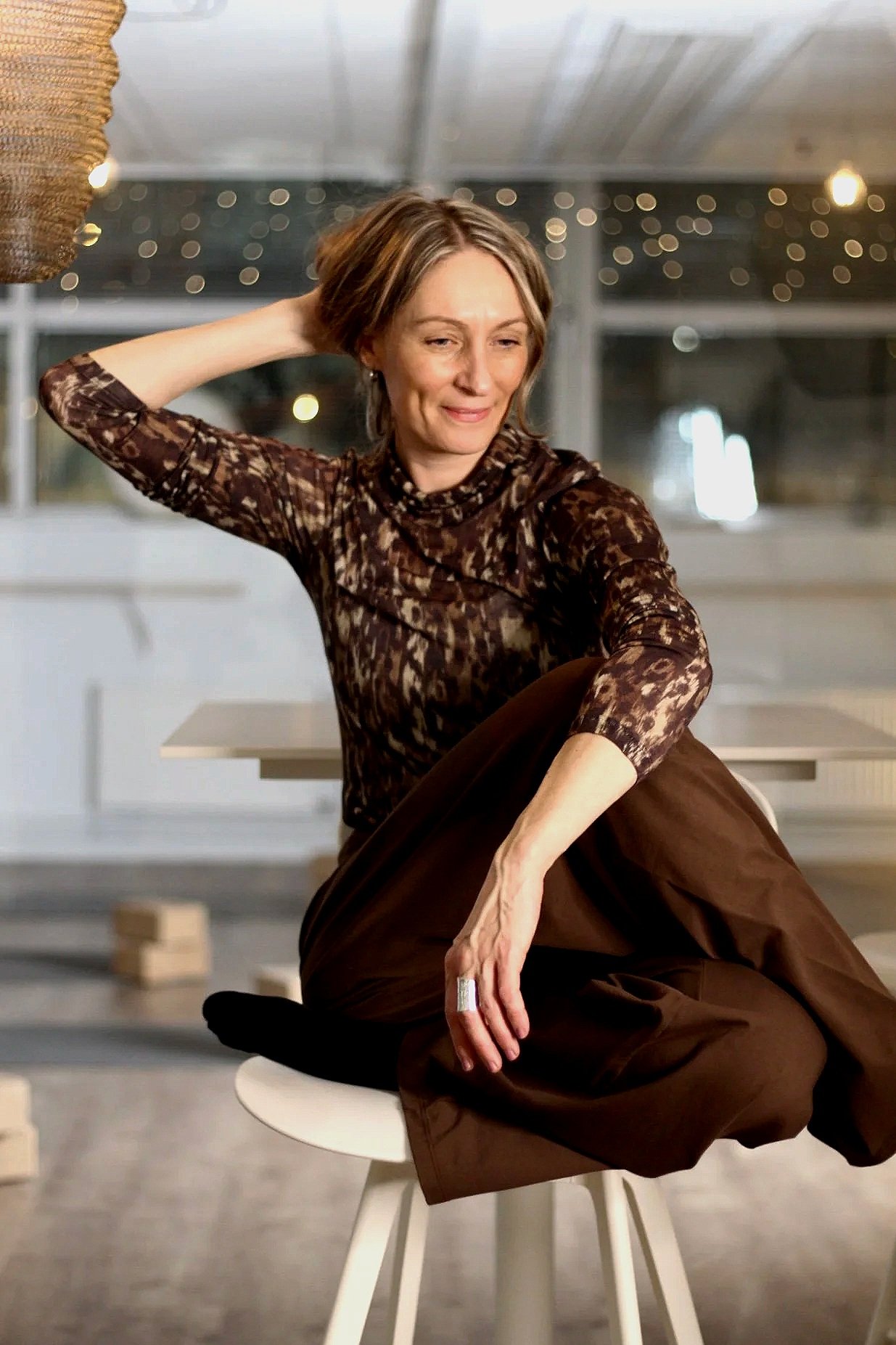 A woman sits on a white stool in a modern indoor space, wearing a brown patterned long sleeve top and brown pants, with her right arm resting on her knee and her left arm behind her head, looking slightly to her left with a smile.