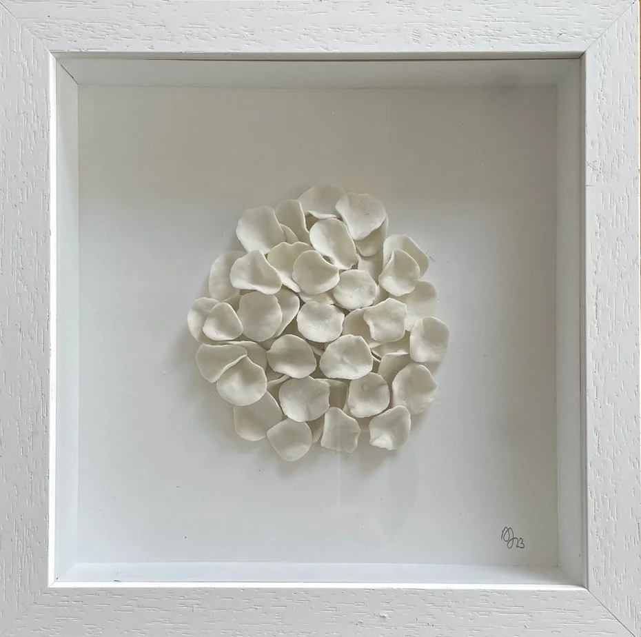A white framed shadow box containing a cluster of white ceramic or porcelain flower petals arranged in a circular pattern.