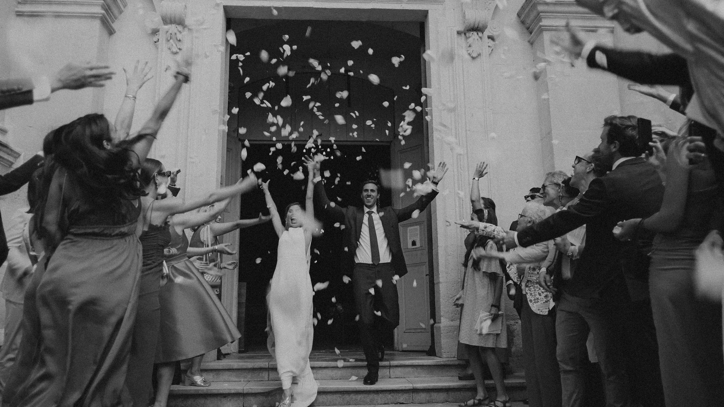 Un couple de mariés joyeux sortant d'une église, entourés d'invités lançant des pétales de fleurs, en noir et blanc.