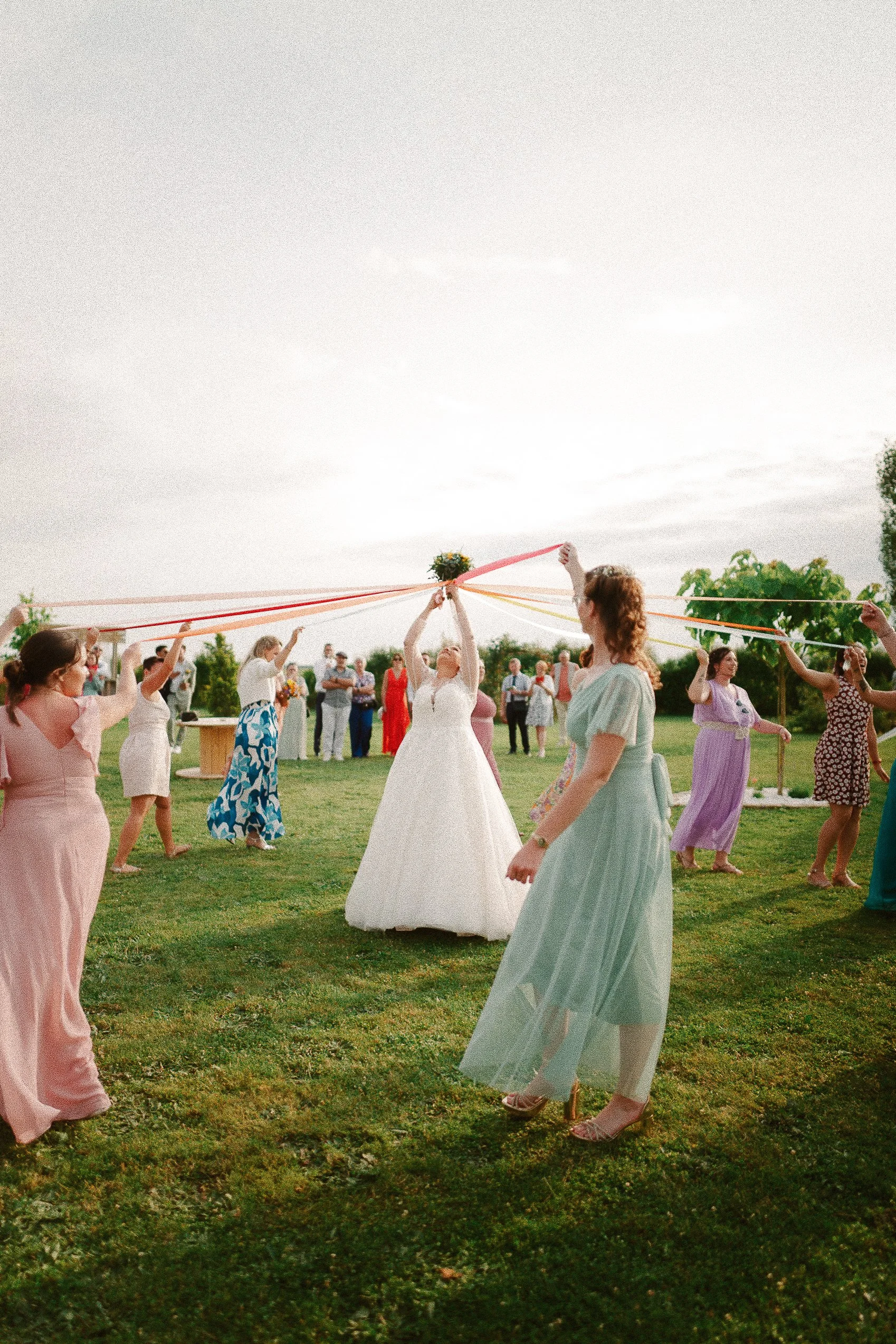 Célébration de mariage en plein air avec la mariée et des invitées formant un cercle, tenant des rubans attachés à un bouquet suspendu au centre, sous un ciel partiellement nuageux.