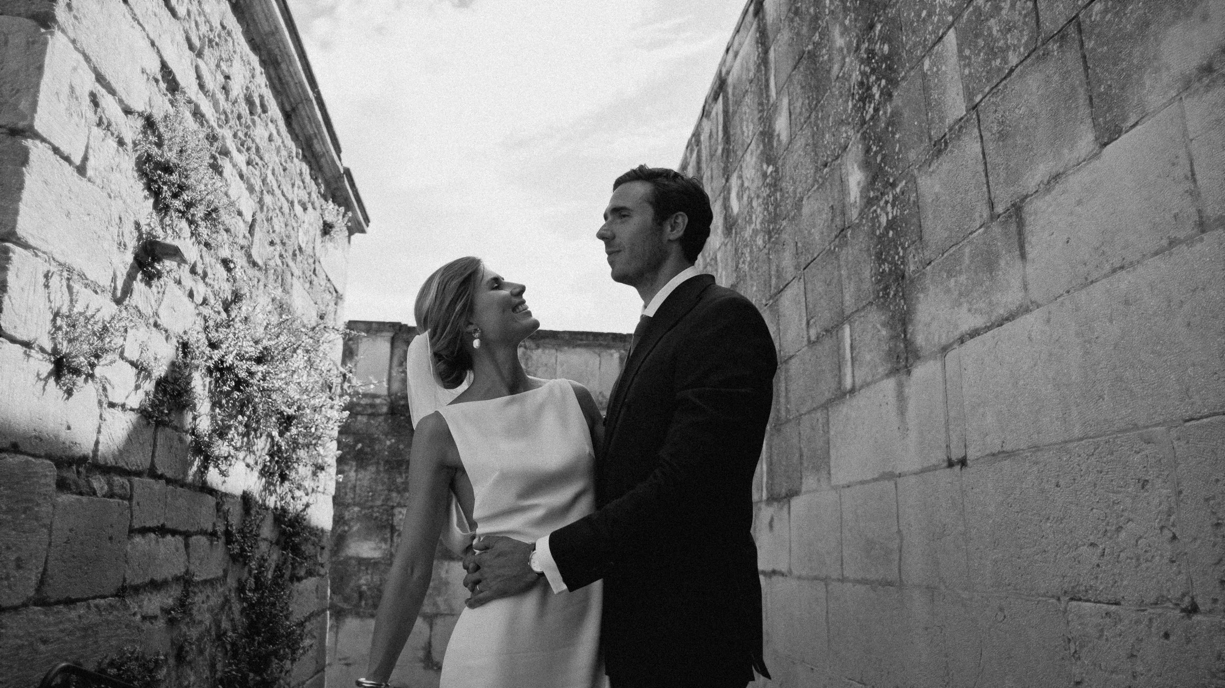 Un couple en tenue de mariage se regarde tendrement dans une ruelle en pierre. La femme sourit, portant une robe blanche et des boucles d'oreilles, tandis que l'homme en costume noir la regarde avec affection.