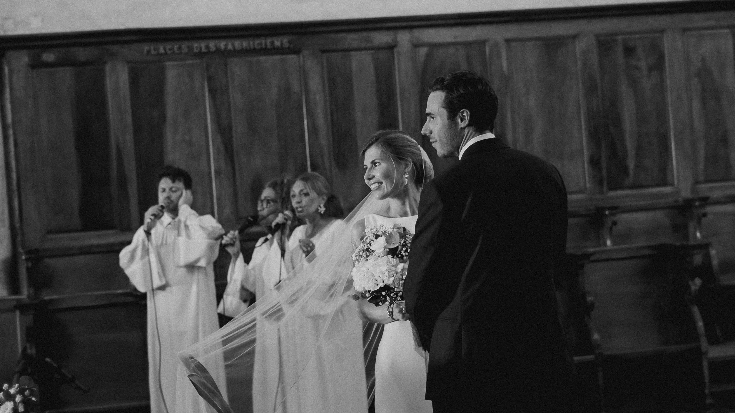 Un couple en mariage échangeant des vœux dans une église, entouré de témoins et d’un pasteur, en noir et blanc.