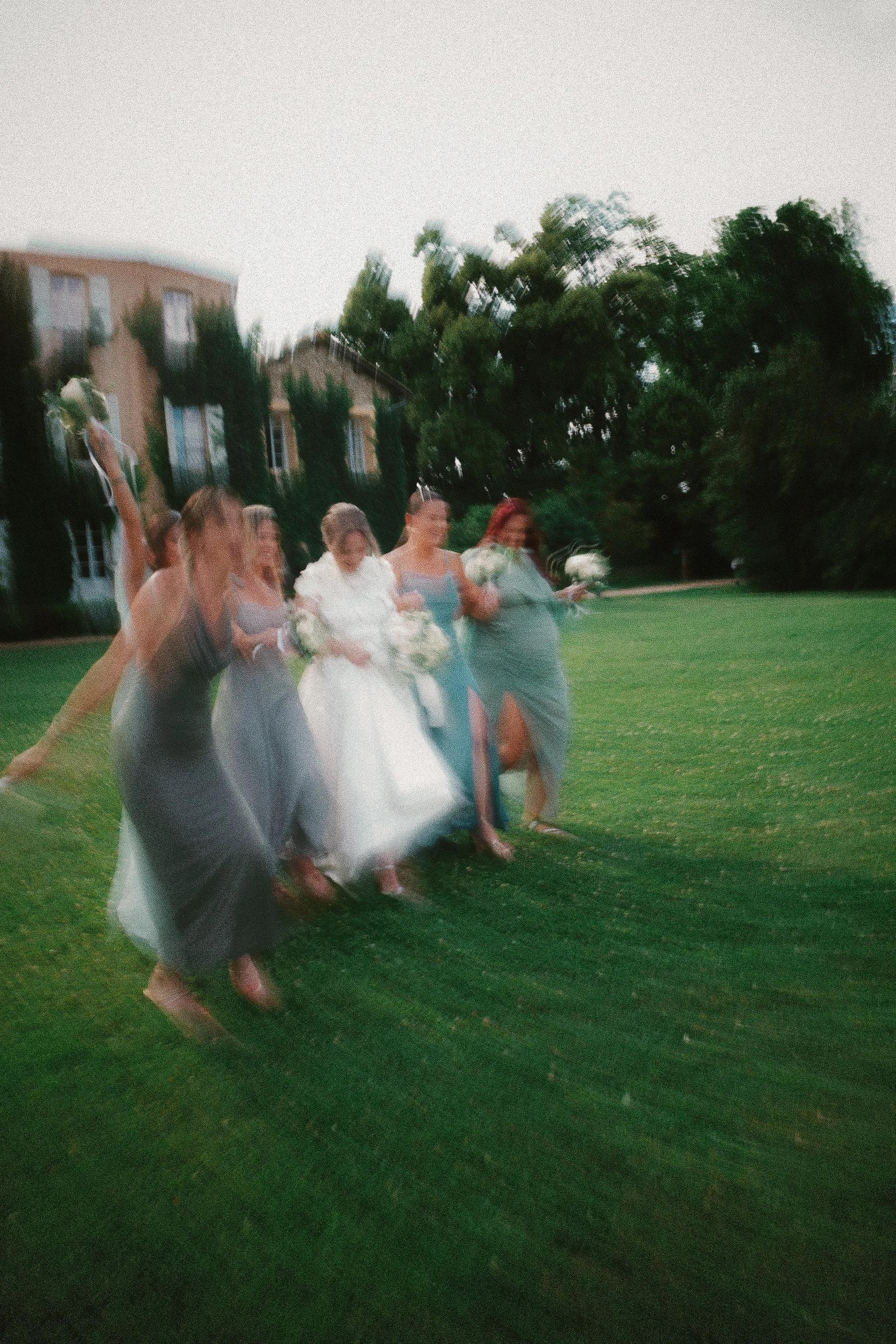 Un groupe de femmes en robes de cérémonie, probablement lors d'un mariage, marchant sur l'herbe d'un parc. La photo semble floue et en mouvement.