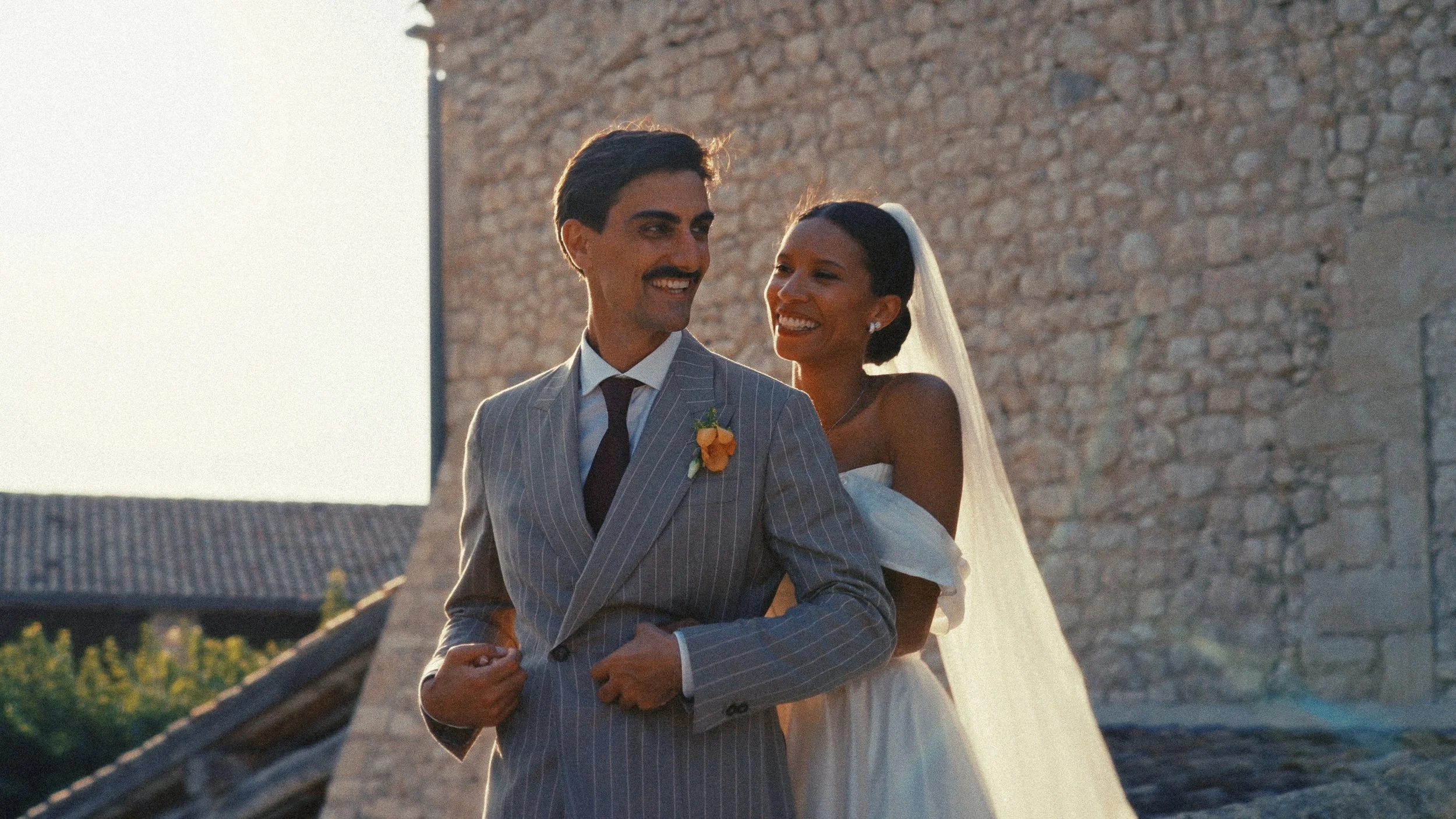 Un couple de mariés souriant, en costume gris rayé et robe blanche, devant un mur en pierre lors d'un mariage en extérieur en soirée.