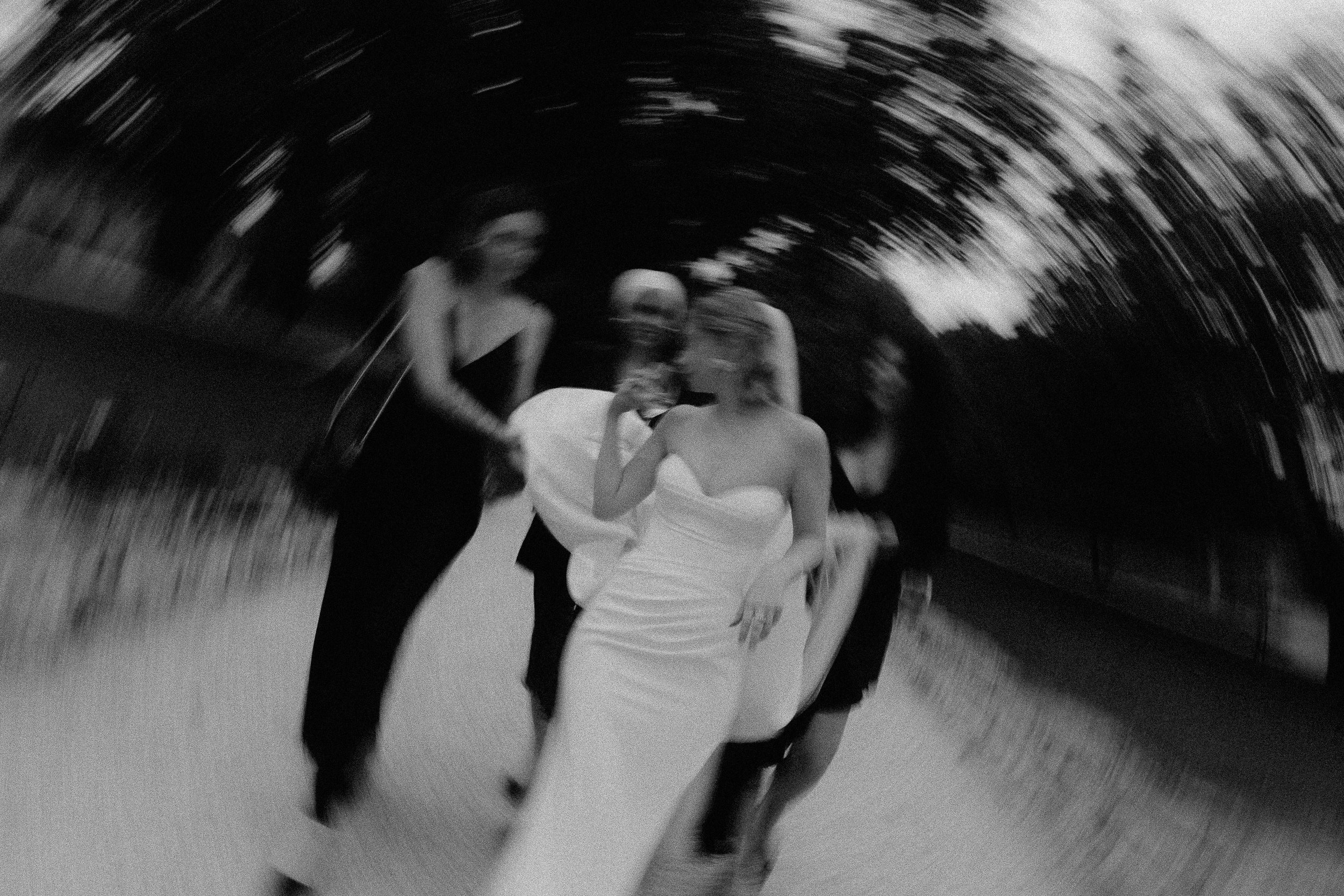 Groupe de femmes debout en conversation, une mariée au milieu, dans un espace extérieur, photographie en noir et blanc avec effet de rotation.