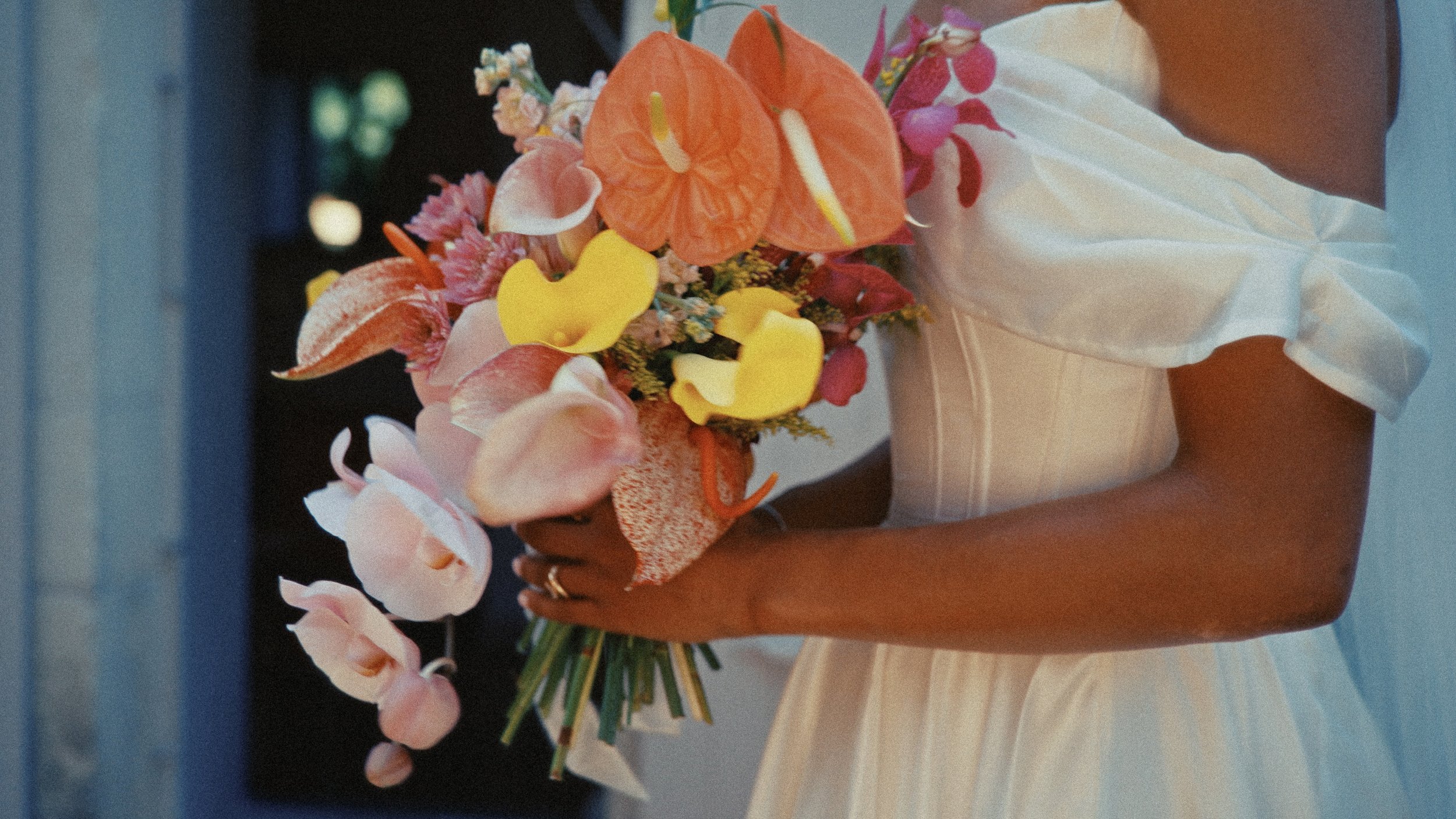 Une personne portant une robe blanche et tenant un bouquet de fleurs colorées, comprenant des callas, des hortensias, des roses, et d'autres fleurs, lors d'une occasion spéciale.