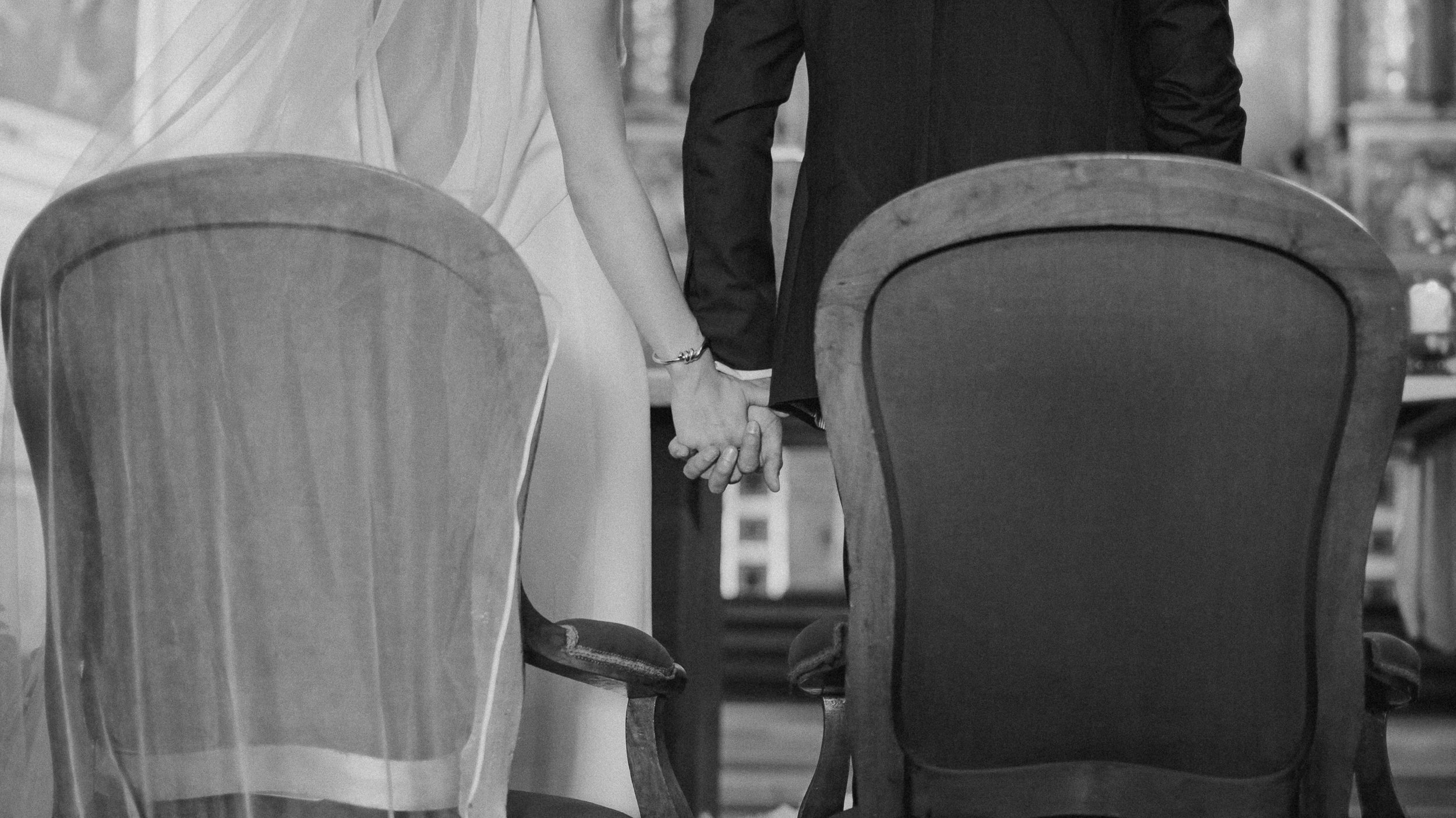 Un couple marié se tenant la main, vu de derrière, assis à une table lors d'une cérémonie, avec toile de voile visible sur la mariée, en noir et blanc.